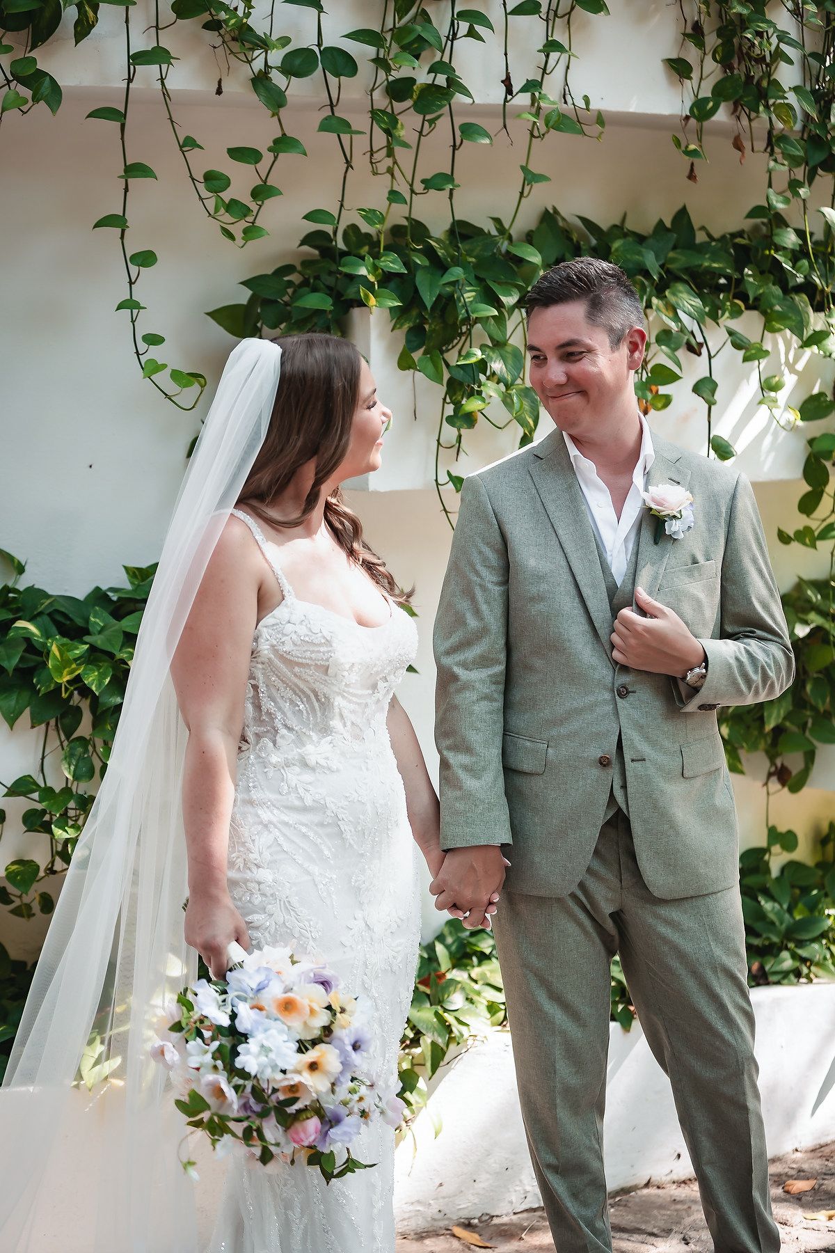 Newlyweds hold hands and smile at each other, bride holding pastel bouquet before ivy backdrop.