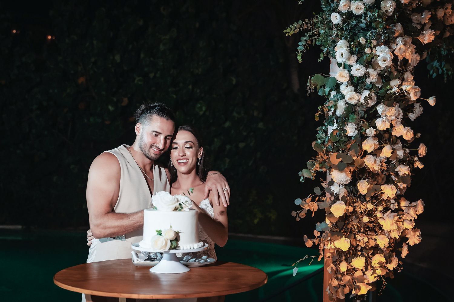 Newlyweds cutting white wedding cake at table beside floral arch at outdoor night reception.