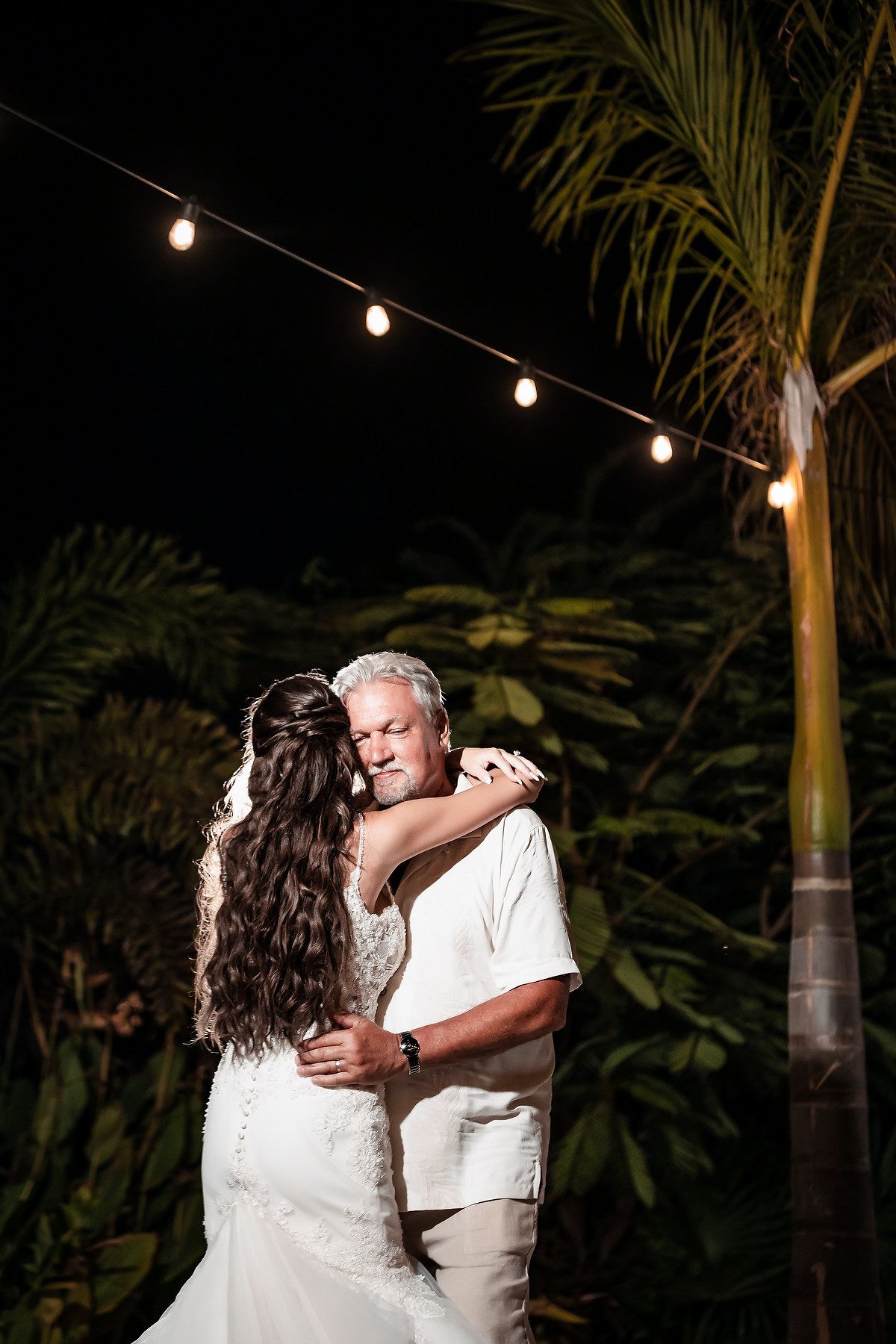 Bride and groom share romantic first dance outdoors at night under string lights with tropical palm trees.