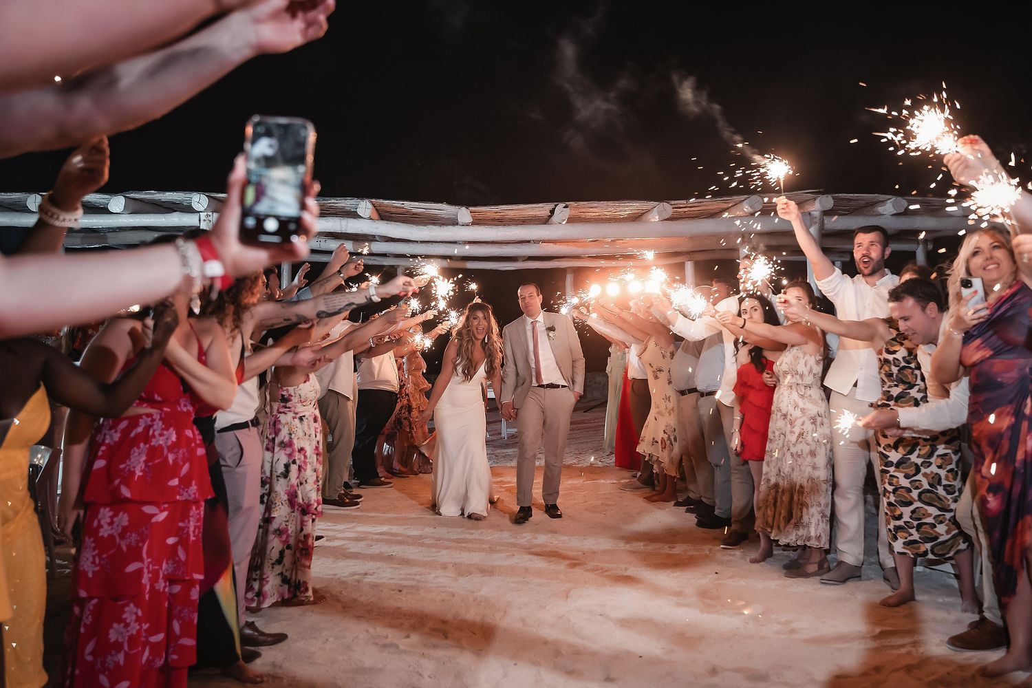 Bride and groom walk through sparkler tunnel surrounded by wedding guests celebrating at night outdoor reception.