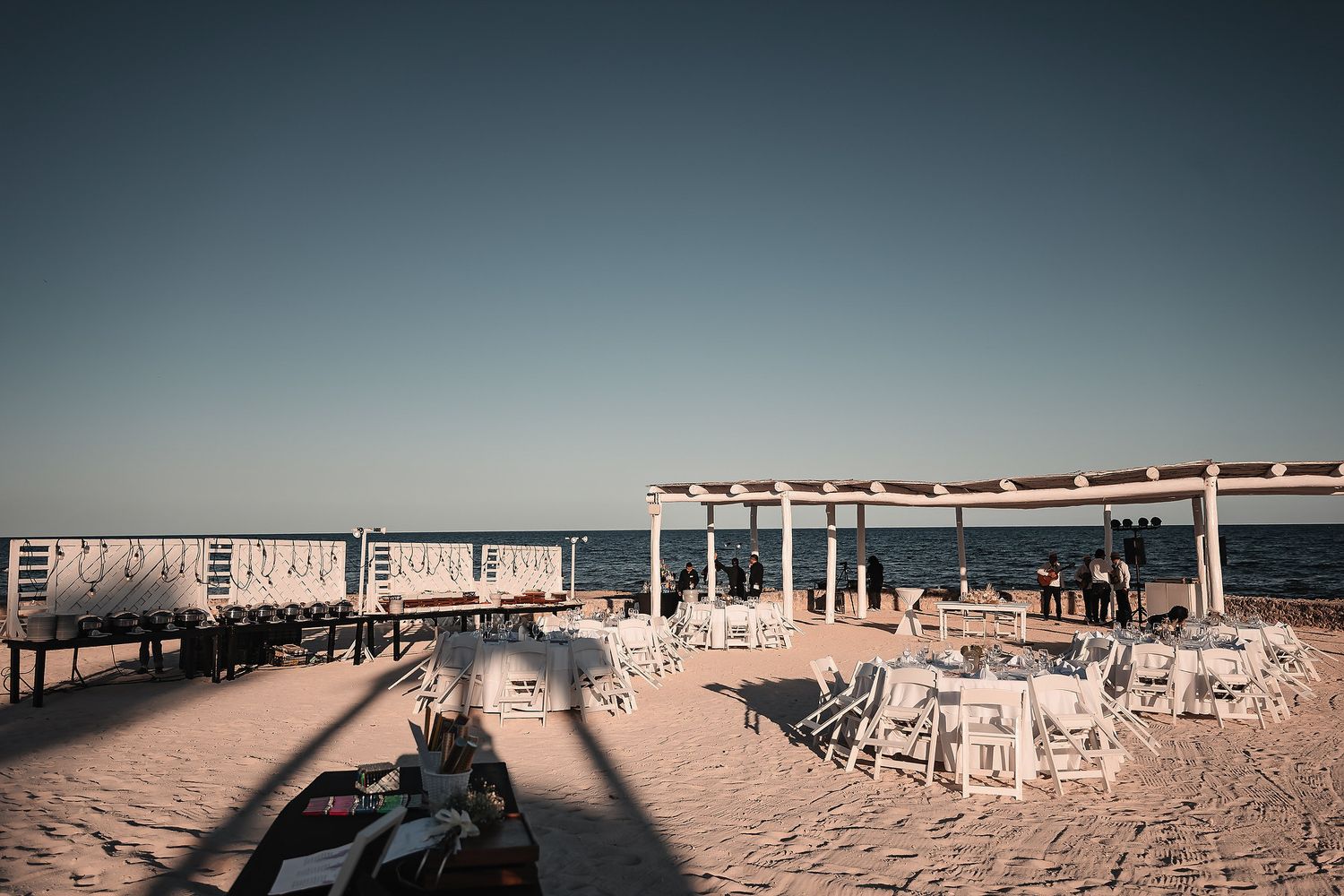 White beach chairs and pergola structure on sandy shore at golden hour with calm ocean backdrop.