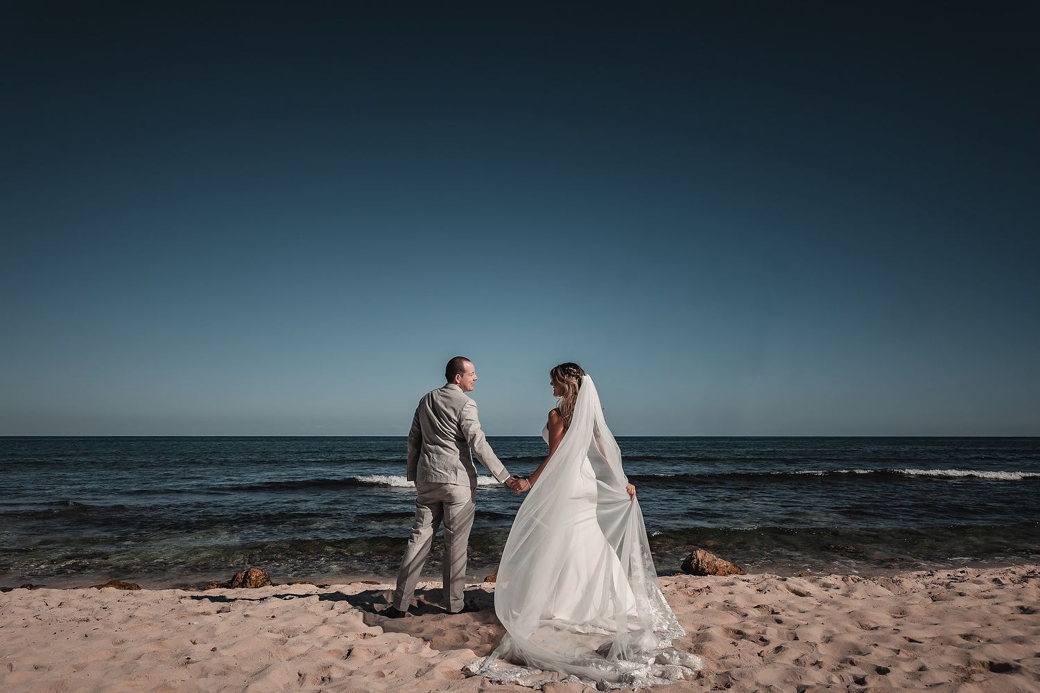 Bride and groom facing each other on a sandy beach with deep blue ocean and clear sky backdrop on their wedding day.