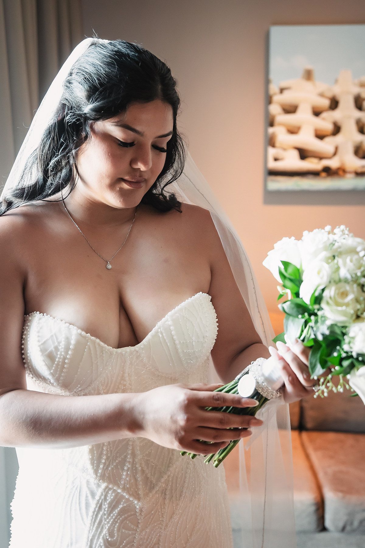 Bride in strapless lace wedding dress with veil holding white rose bouquet indoors.