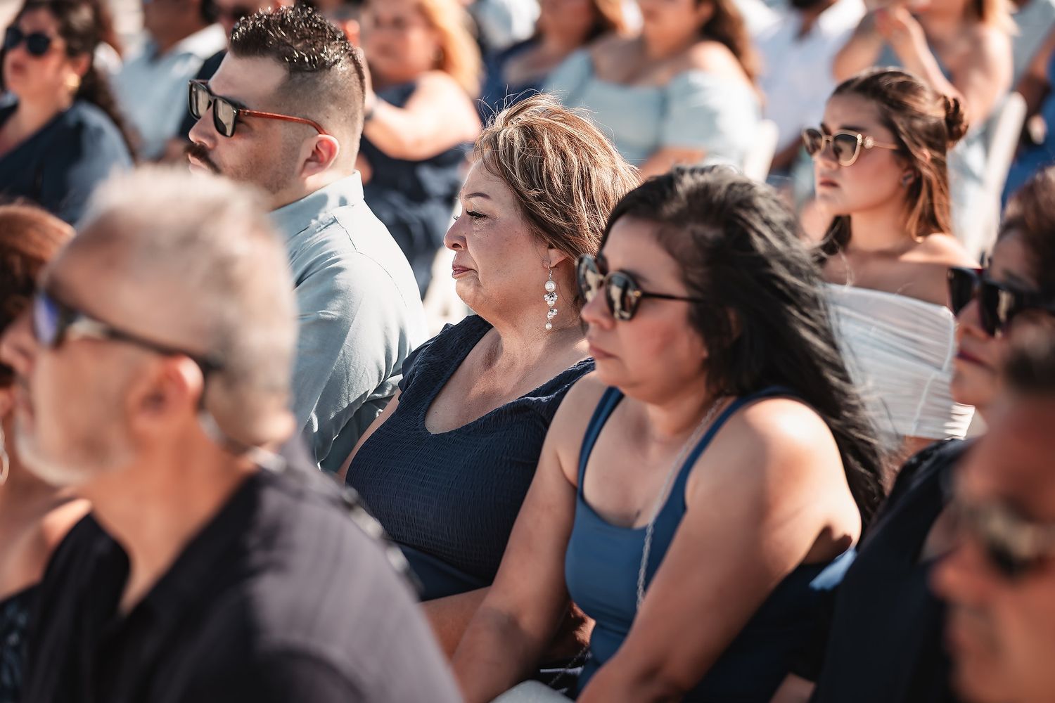 Crowd of diverse people sitting outdoors at a sunny event, wearing casual attire and sunglasses.