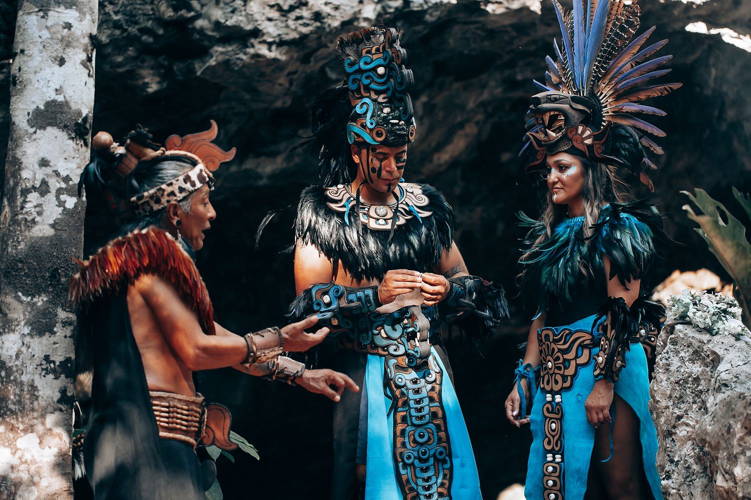 Three performers in detailed Aztec ceremonial dress with turquoise and feathered headdresses converse near a cave entrance.