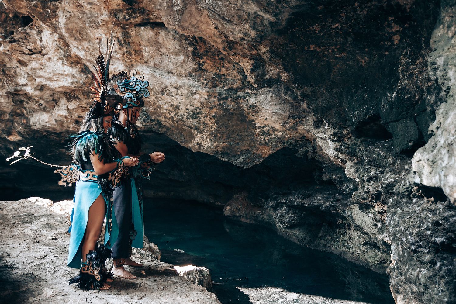 Two people in Mayan ceremonial costumes with feathered headdresses stand together inside a rocky cave entrance.