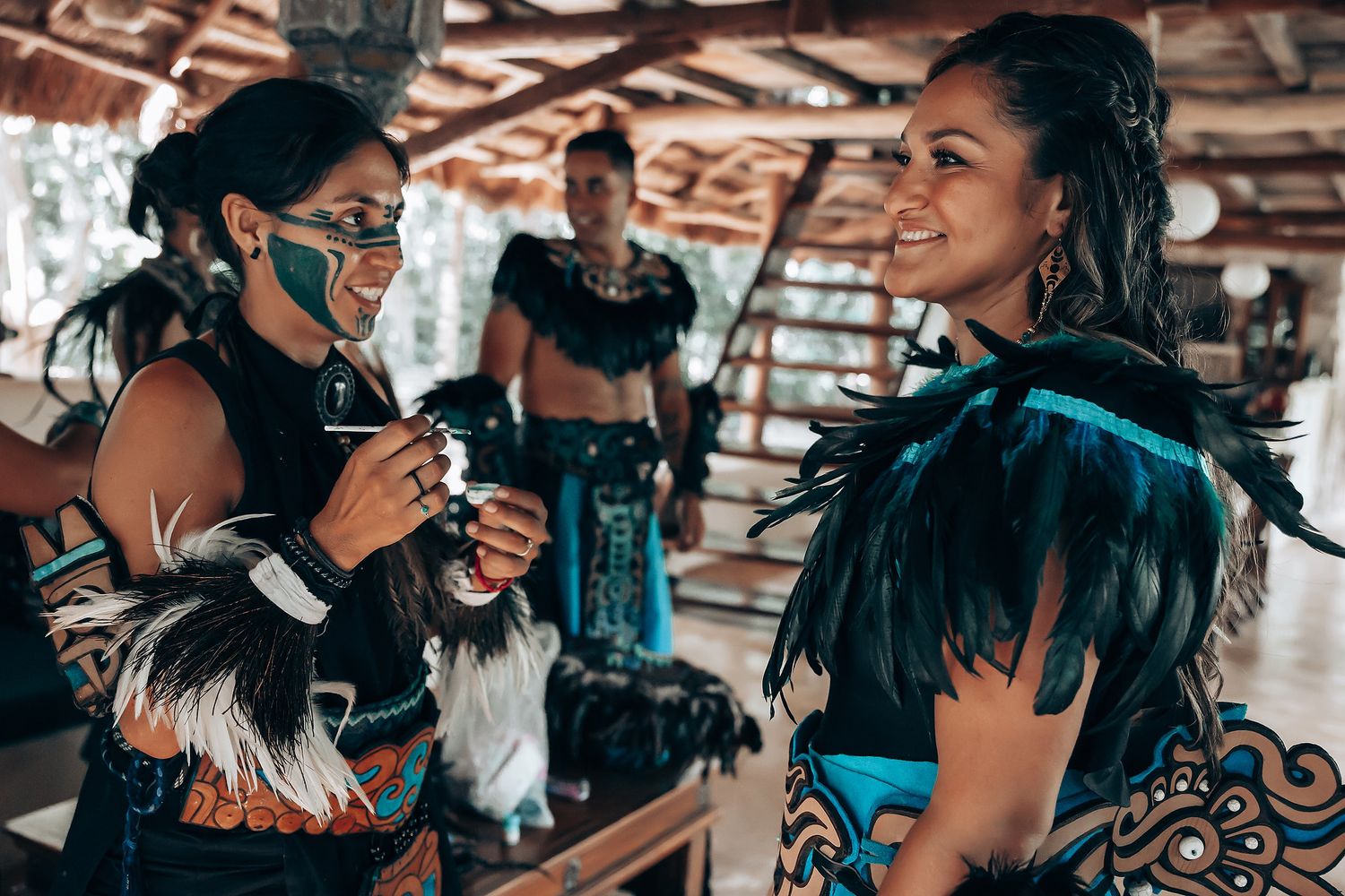 Two women in turquoise and black Mayan ceremonial feathered costumes conversing indoors.