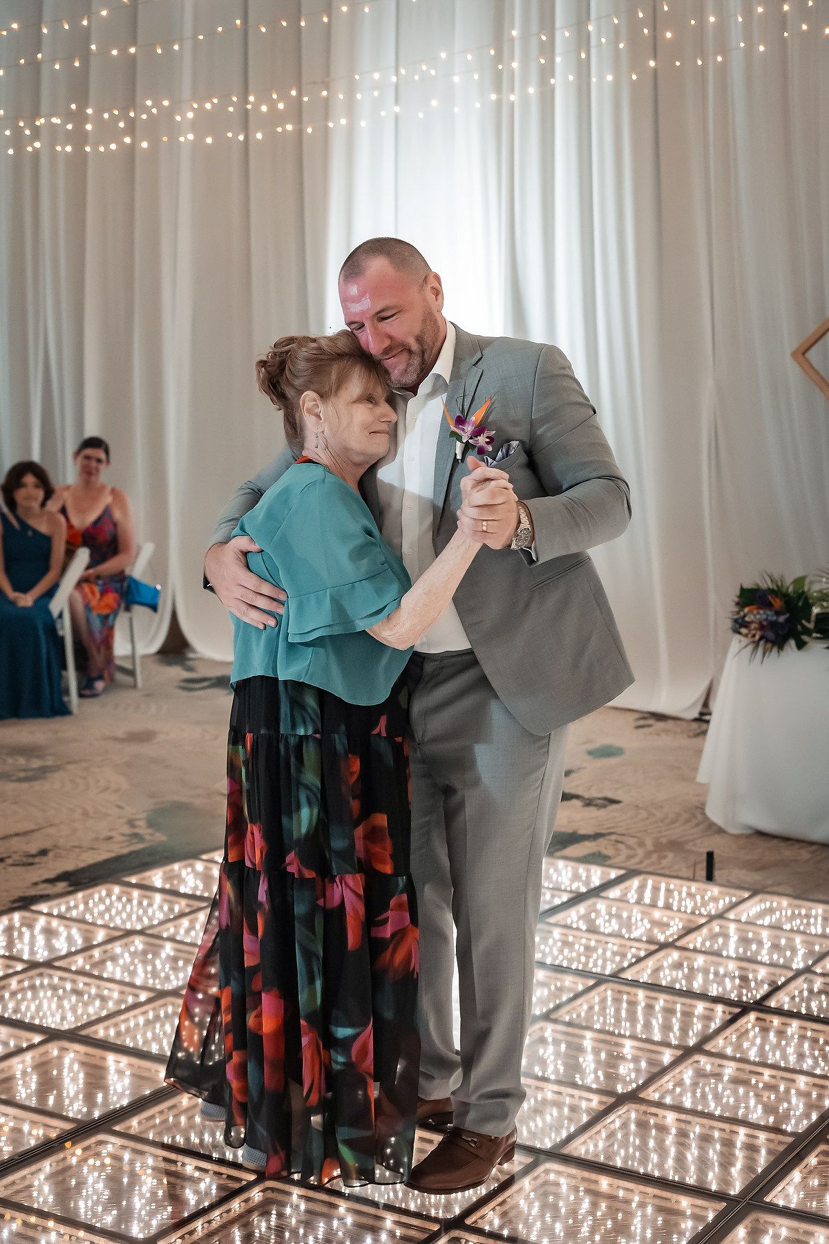 Groom in gray suit shares emotional mother-son dance on sparkling LED dance floor at elegant wedding reception.