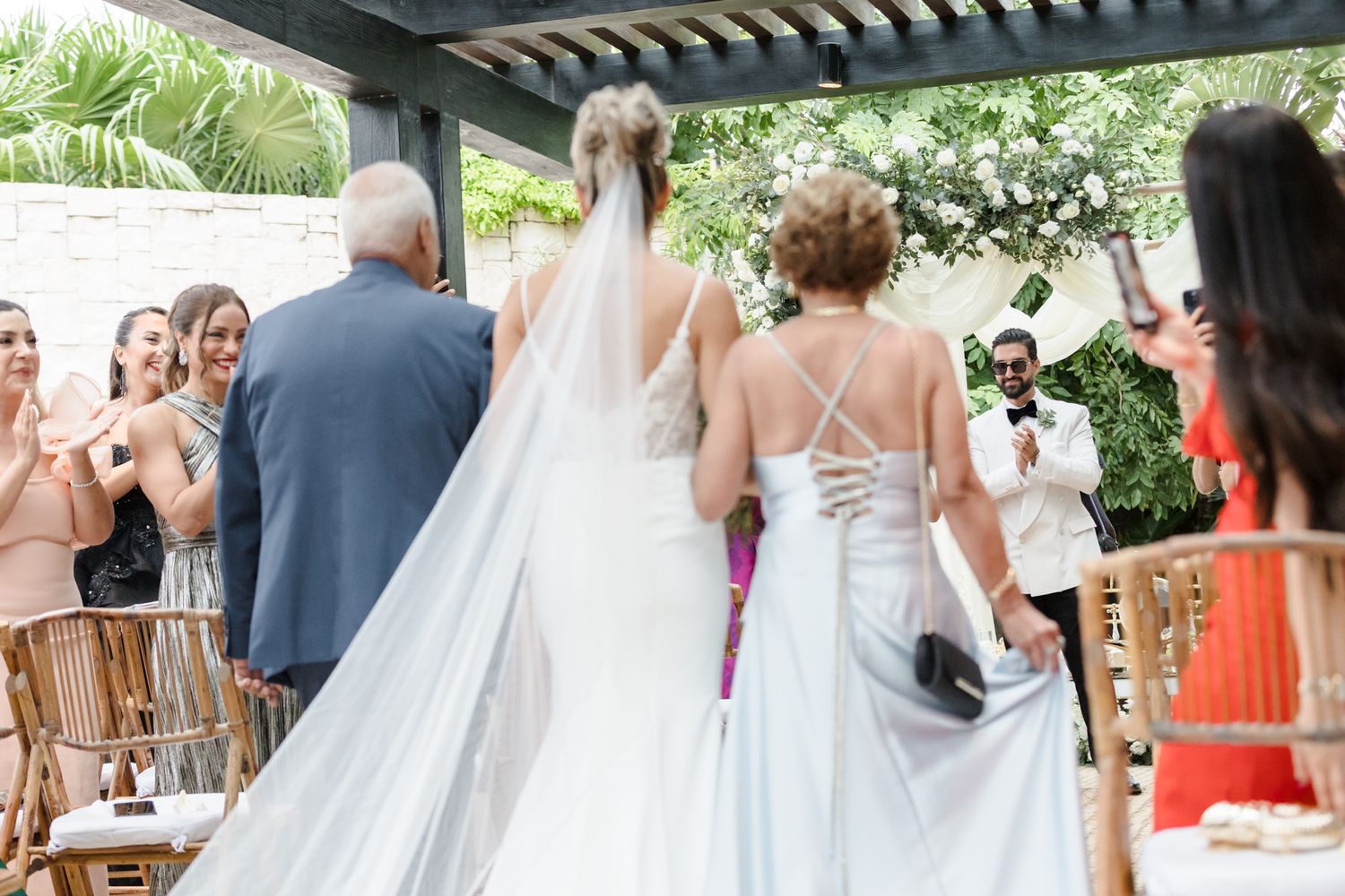 Bride in white gown with long veil walks toward altar at an outdoor wedding ceremony.