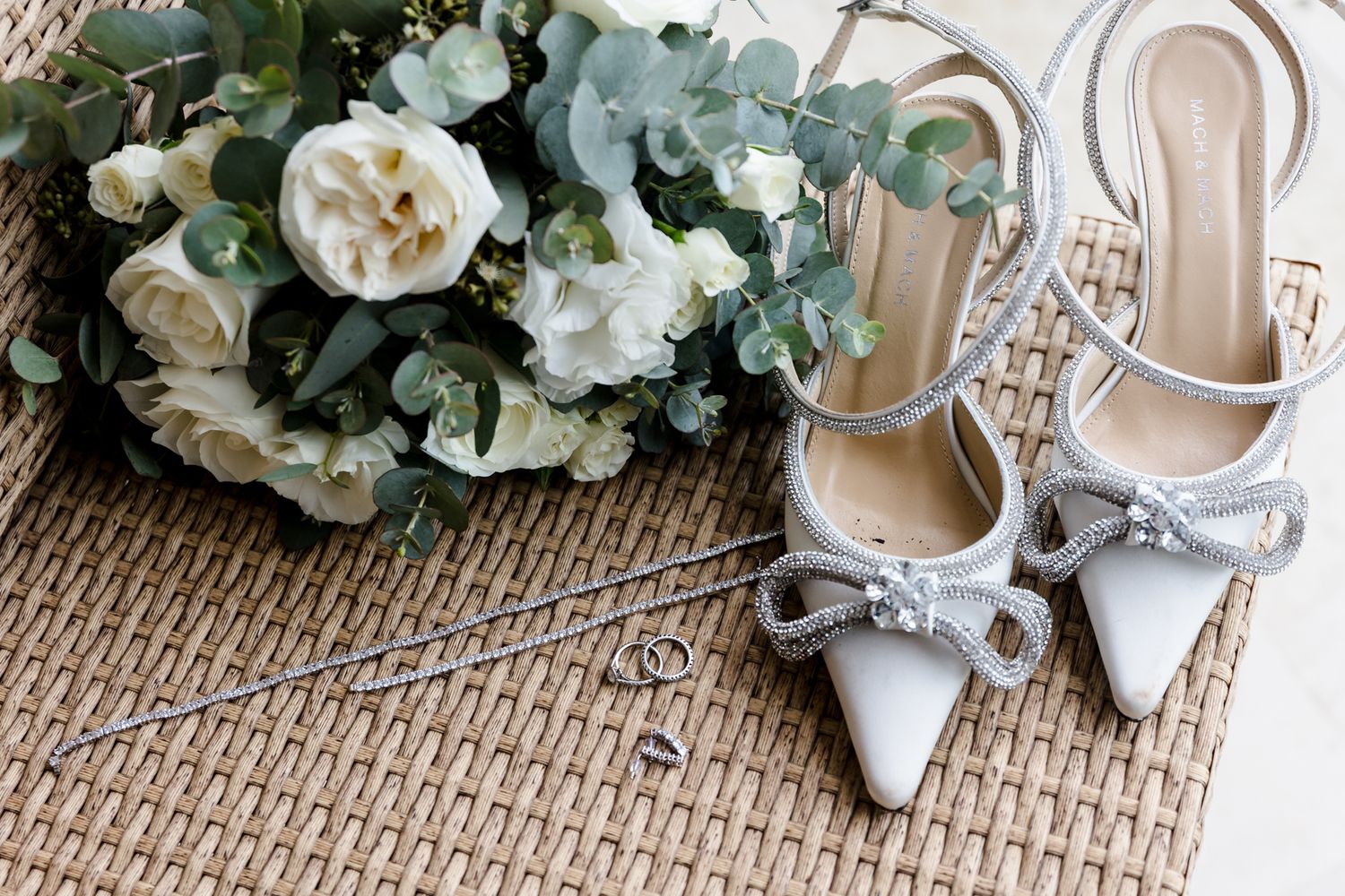 Elegant bridal accessories including white heels with bow detail, silver earrings, rings, and a white floral bouquet on wicker.