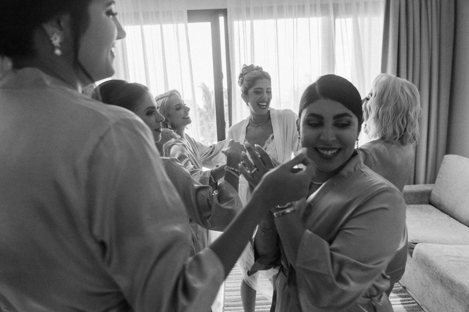 Black and white photo of a bride getting ready with bridesmaids in robes, laughing and sharing a joyful moment.