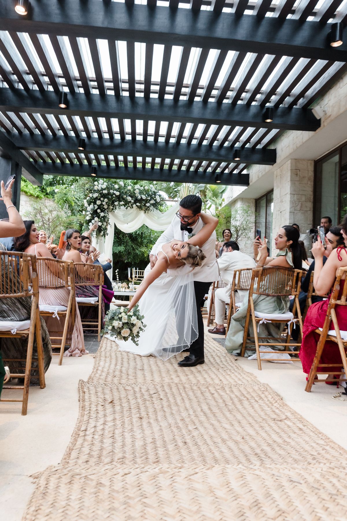 Groom dips bride in wedding aisle after ceremony, guests cheering, floral arch backdrop, outdoor venue with pergola.