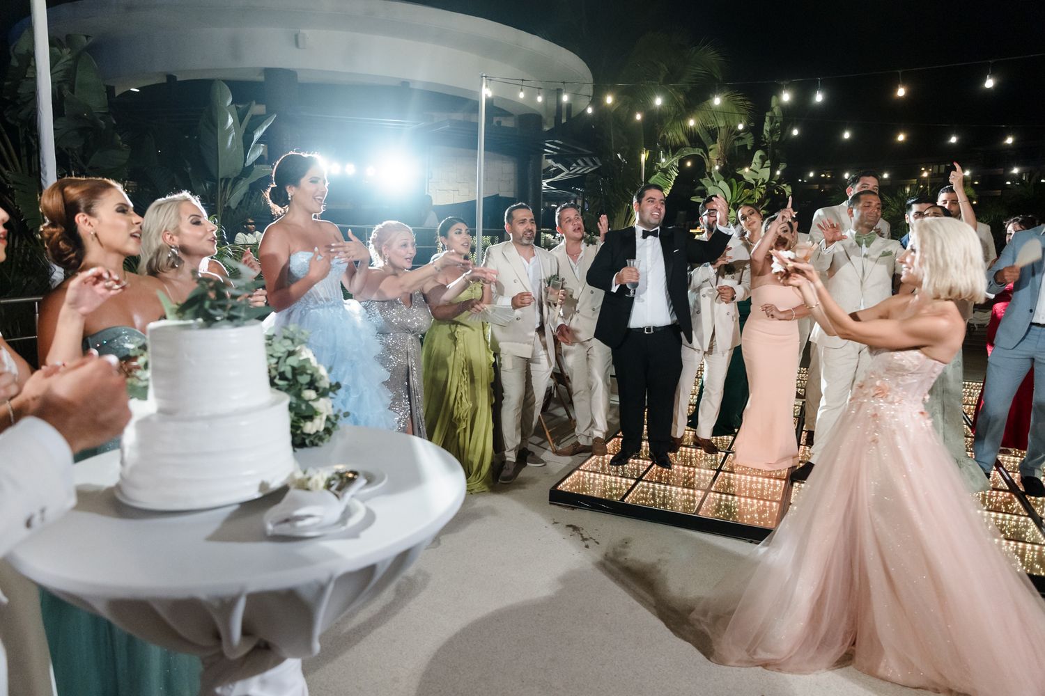 Elegant outdoor wedding reception with guests celebrating, a white tiered cake, and string lights at night.