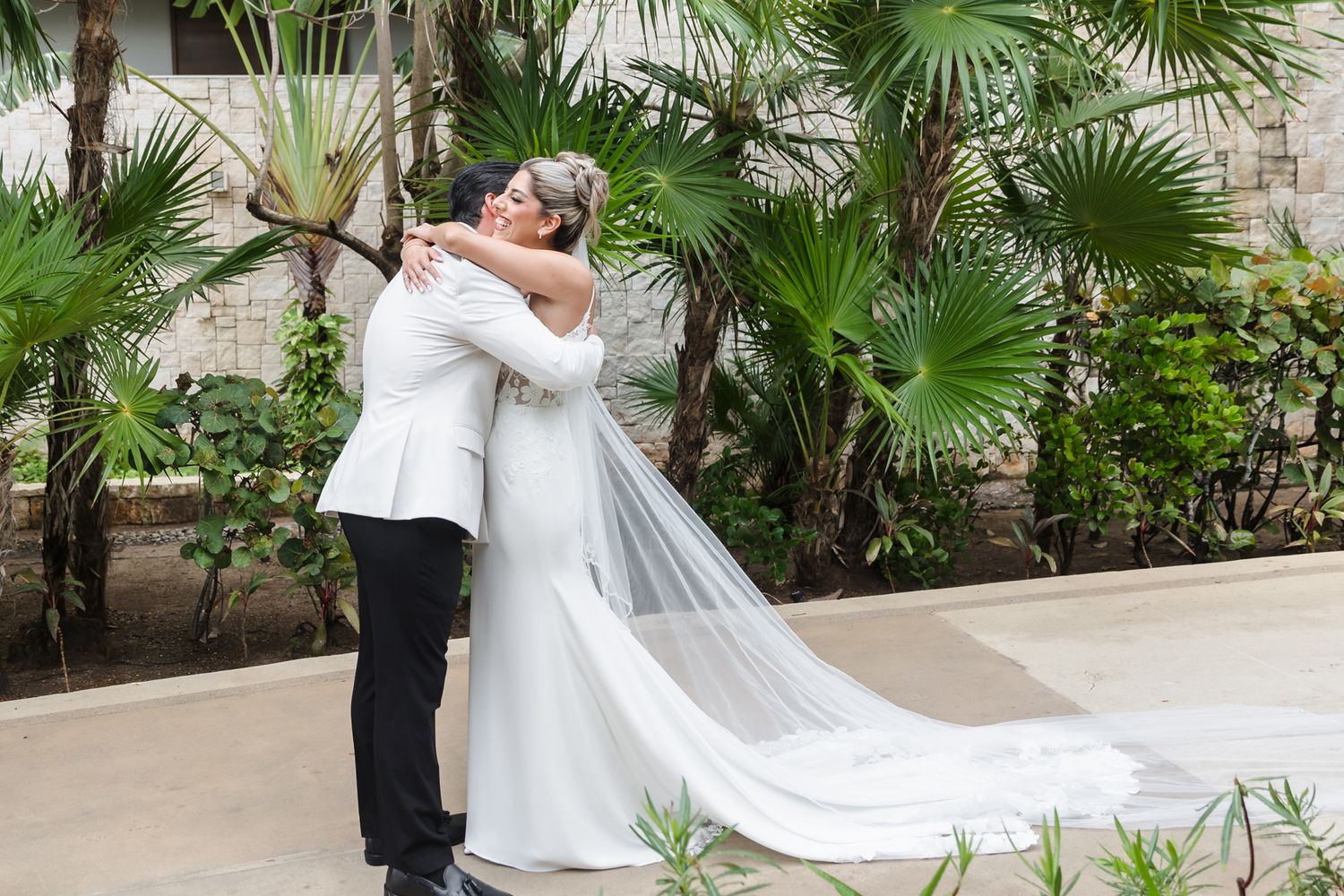 Bride and groom share a romantic embrace outdoors, surrounded by lush tropical palm trees, as her veil flows gracefully.
