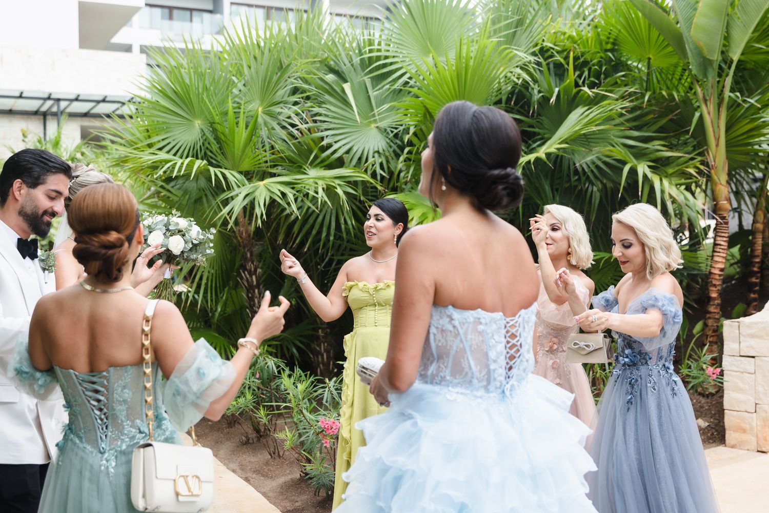 Women in colorful ball gowns gather outdoors near tropical palm trees at an elegant garden party or prom event.