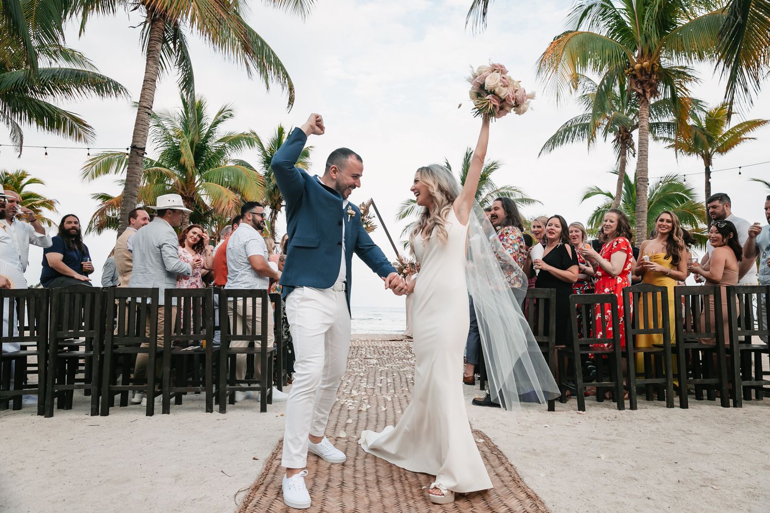 Joyful newlyweds raise hands celebrating after beach wedding ceremony surrounded by palm trees and cheering guests.