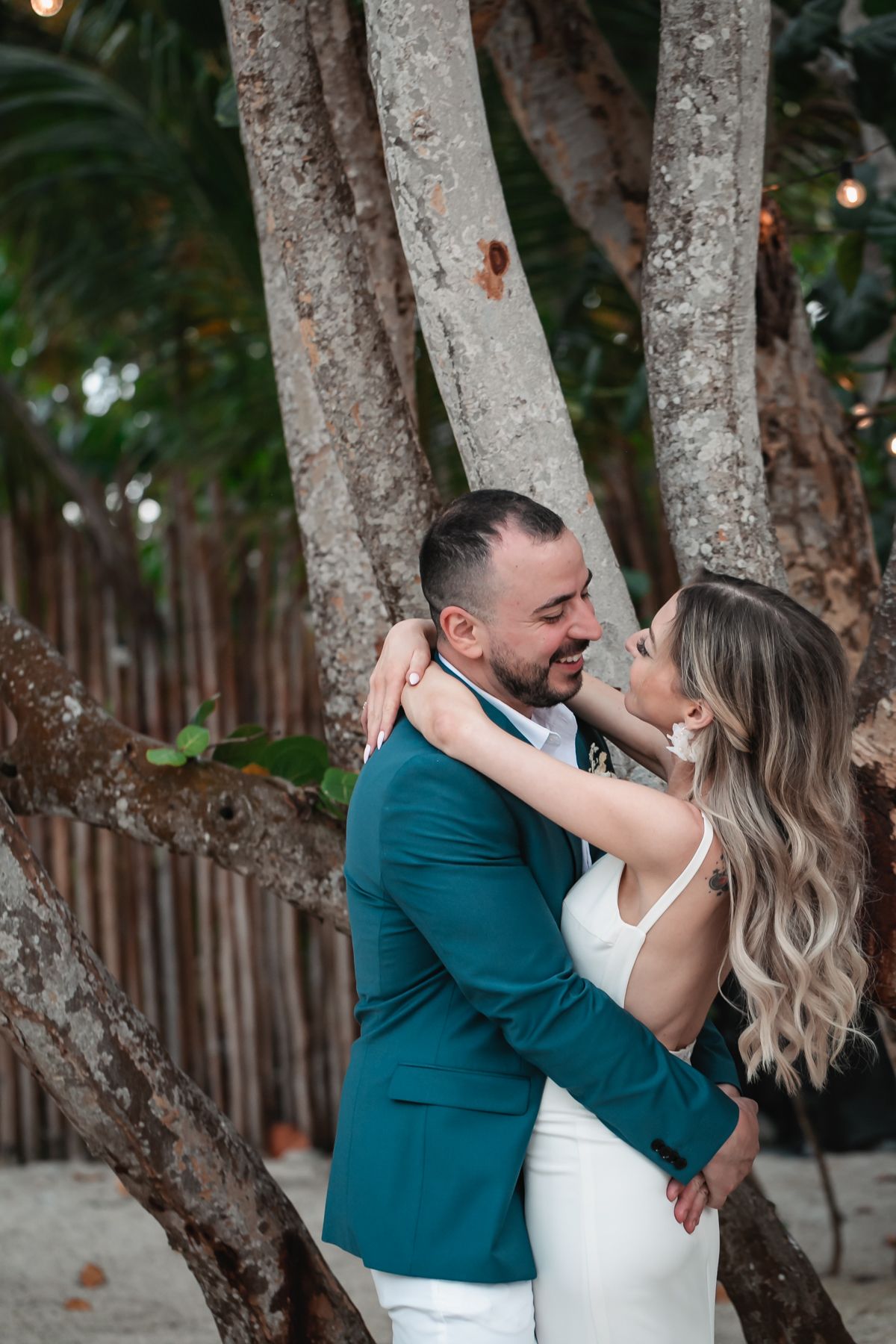 Bride and groom share romantic moment embracing among tropical trees, groom in teal blazer and bride in white gown.