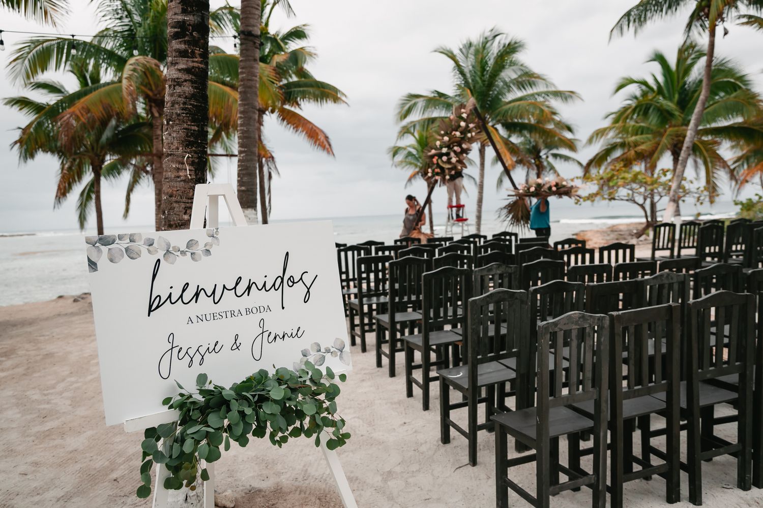 Beach wedding ceremony setup with black chairs, palm trees, and a bienvenidos welcome sign for Jessie & Steve.