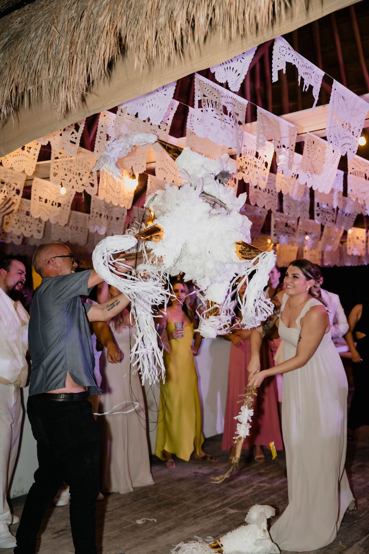 Bride smashing white piñata at colorful tropical wedding reception decorated with papel picado.