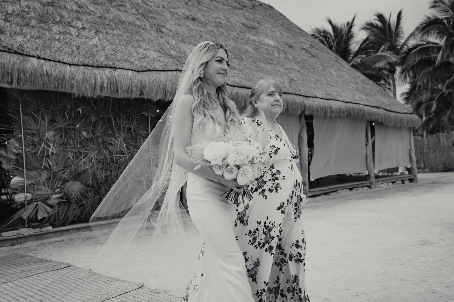 Black and white photo of bride in veil and gown walking with mother near thatched huts.