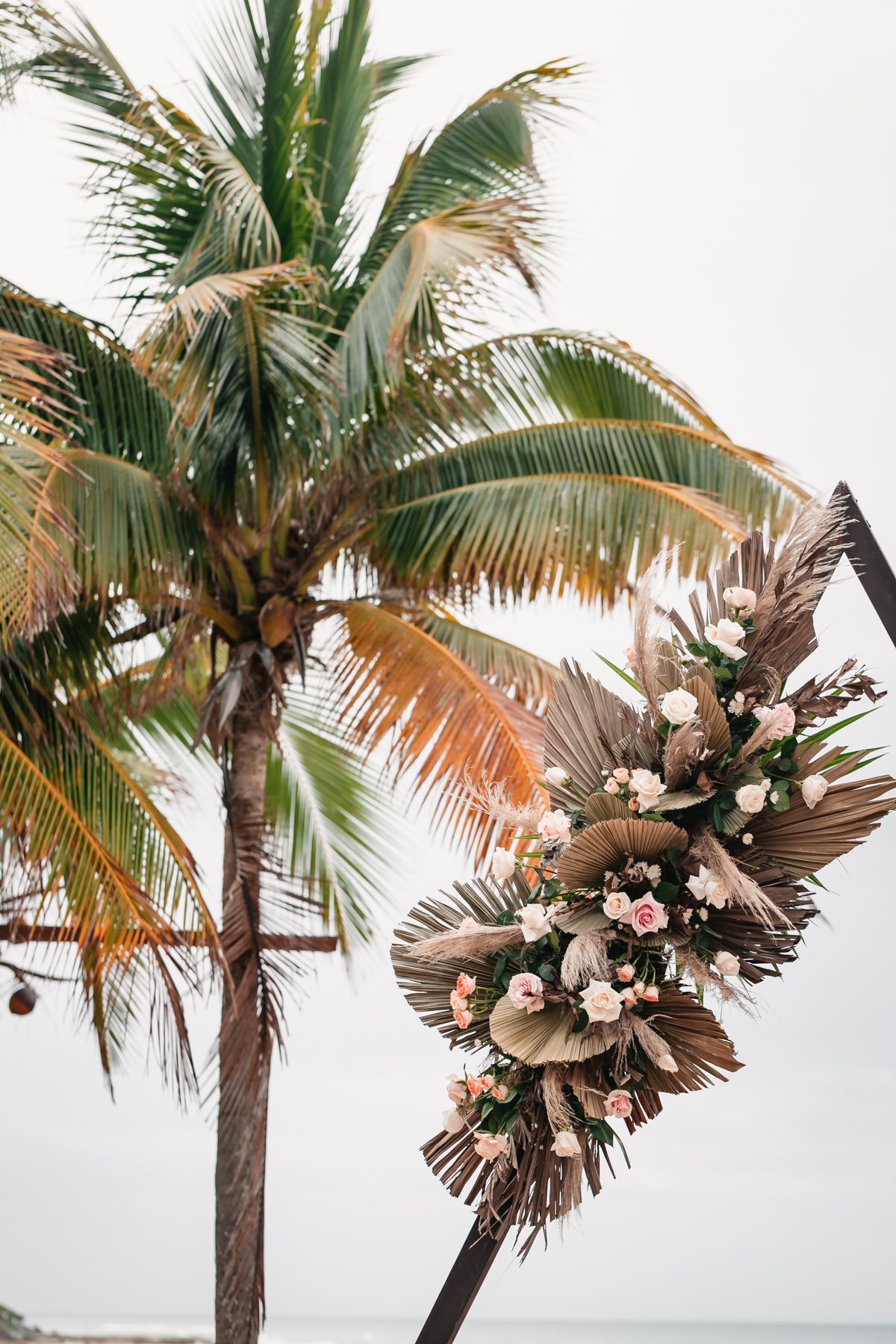 Tropical beach wedding arch adorned with pink roses and dried palm leaves with a coconut palm tree backdrop.