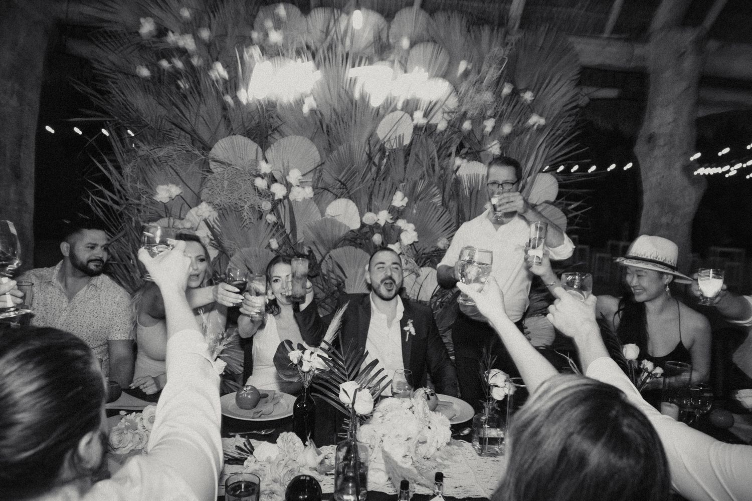 Black and white photo of wedding guests raising glasses in toast at tropical reception dinner.