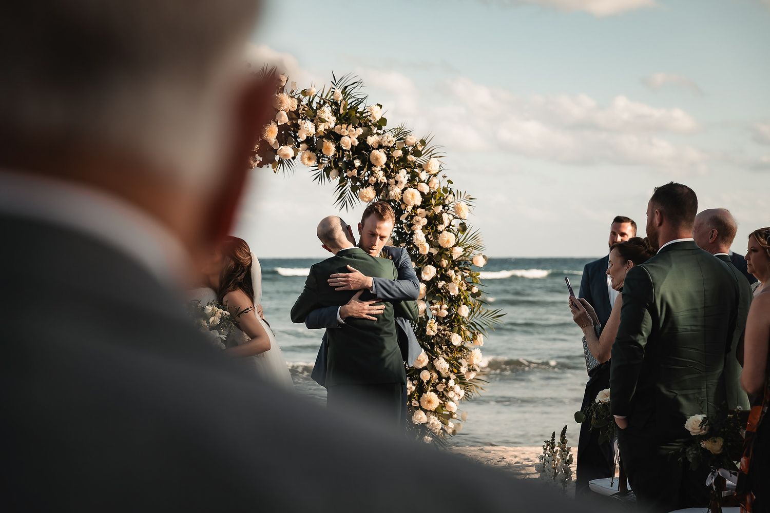 Couple embracing at a beach wedding ceremony with a floral arch backdrop and ocean views.
