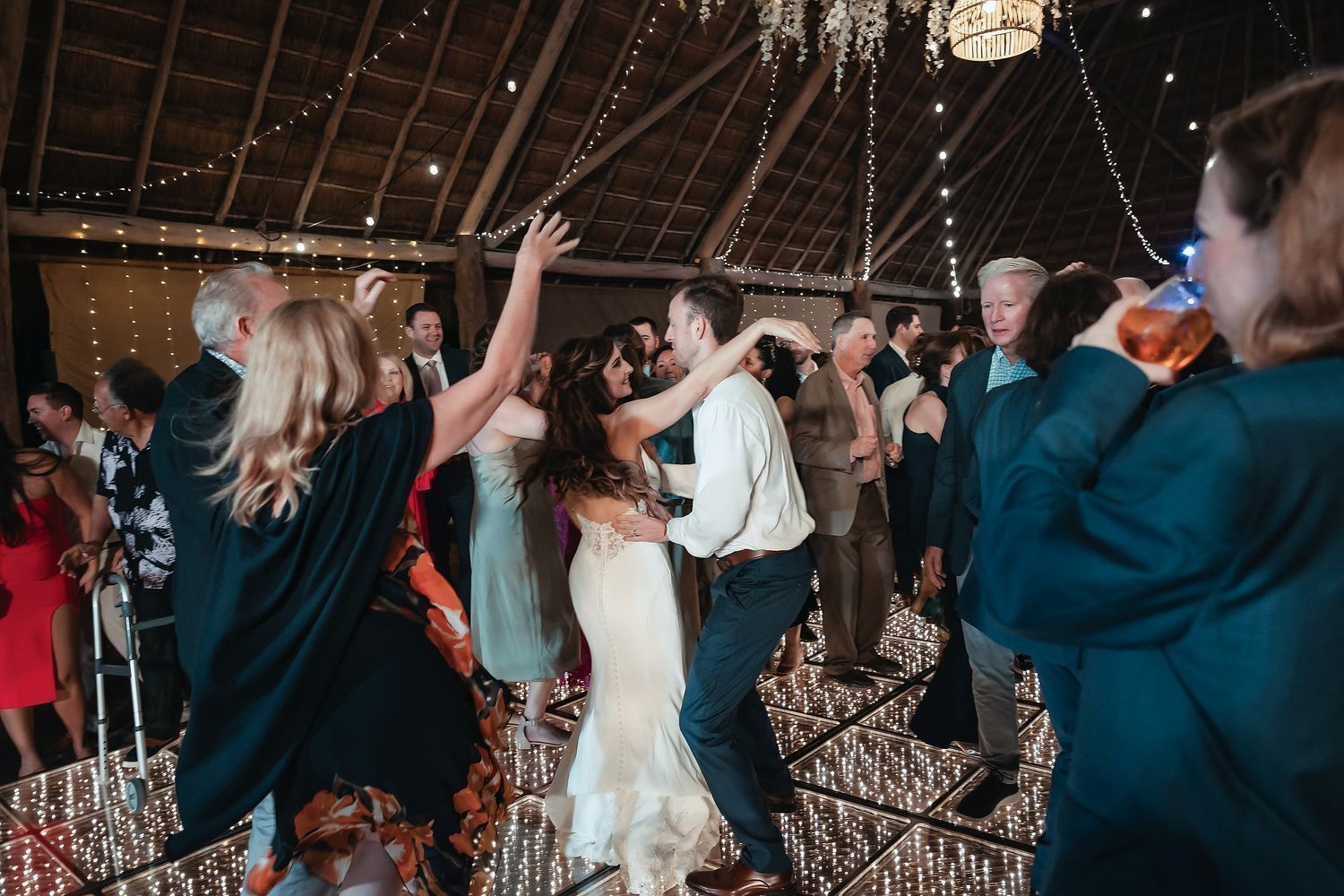 Bride and groom dancing joyfully with guests at a wedding reception inside a decorated barn venue.