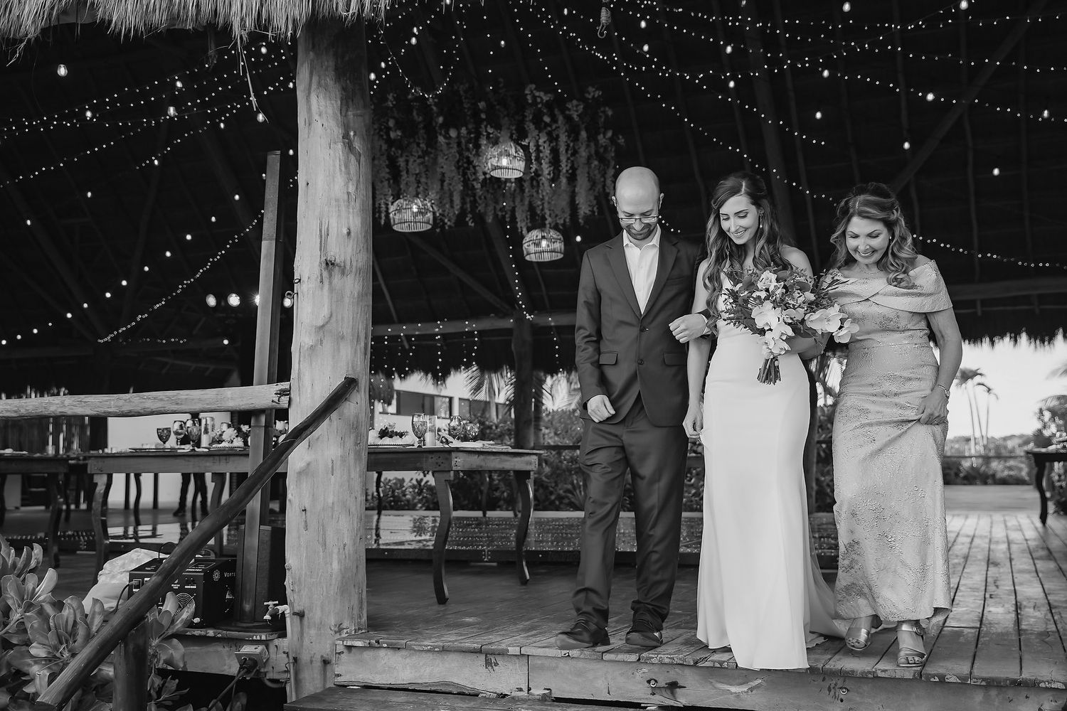 Black and white photo of bride with bouquet escorted by two people at rustic beach wedding.