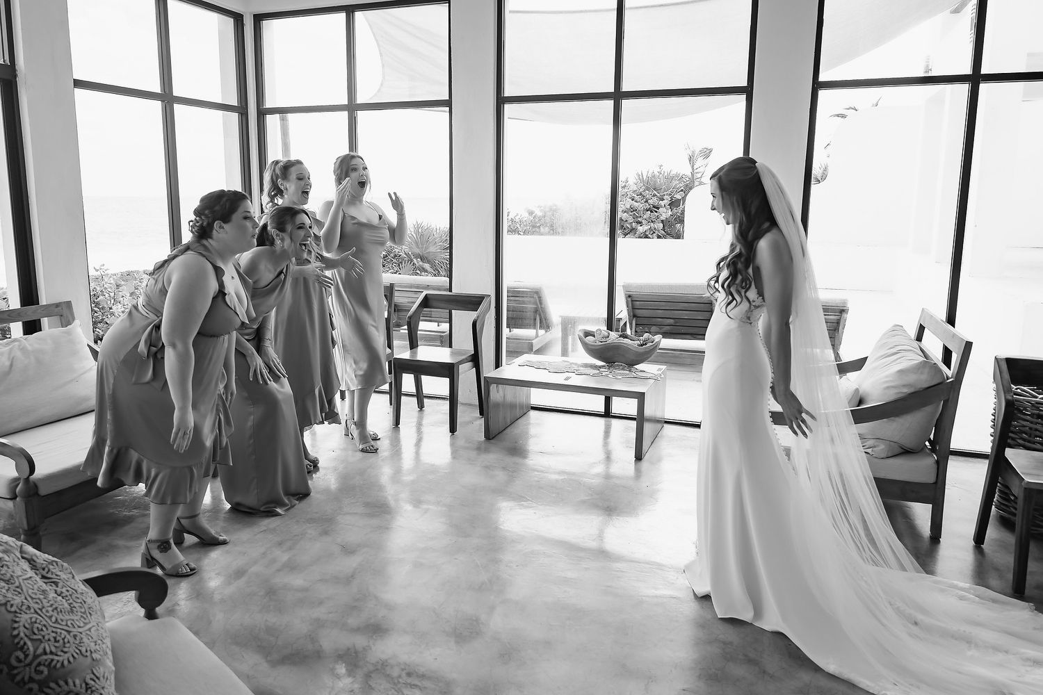 Bride in white gown reveals look to bridesmaids in gray dresses in a sunlit room during wedding preparations.