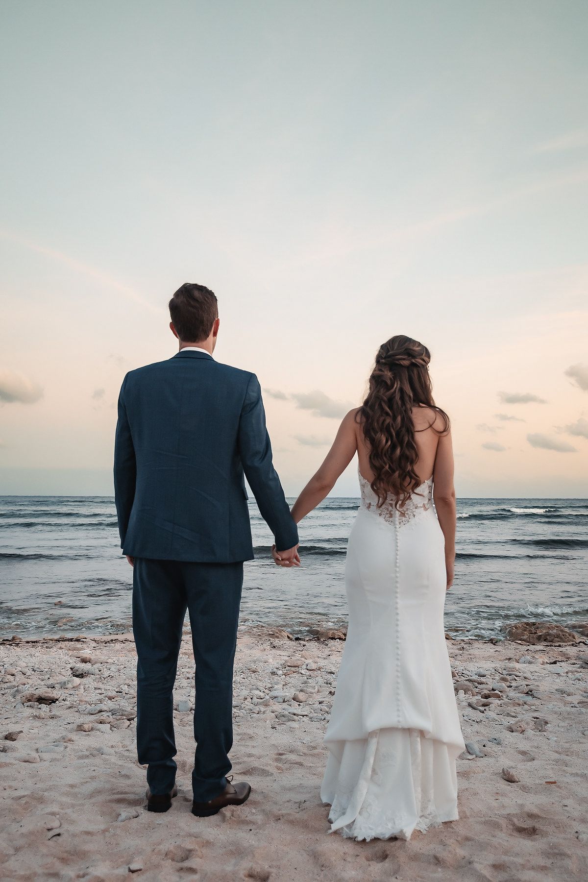 Bride and groom holding hands on a sandy beach at sunset, viewed from behind, wearing wedding attire.