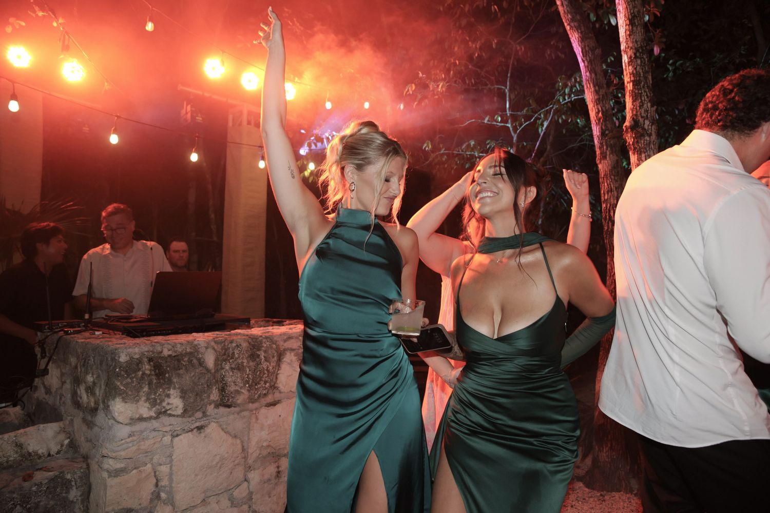 Two women in emerald green satin gowns dancing at an outdoor evening wedding reception with dramatic red lighting.