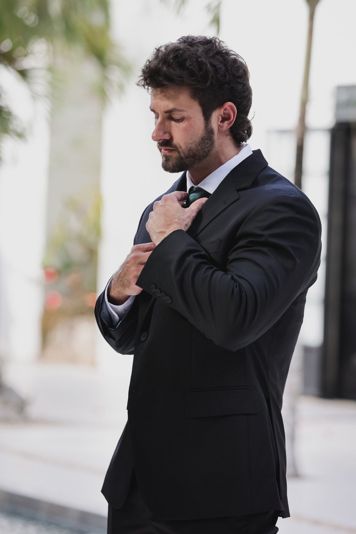 A well-dressed man in a black suit adjusting his tie outdoors.