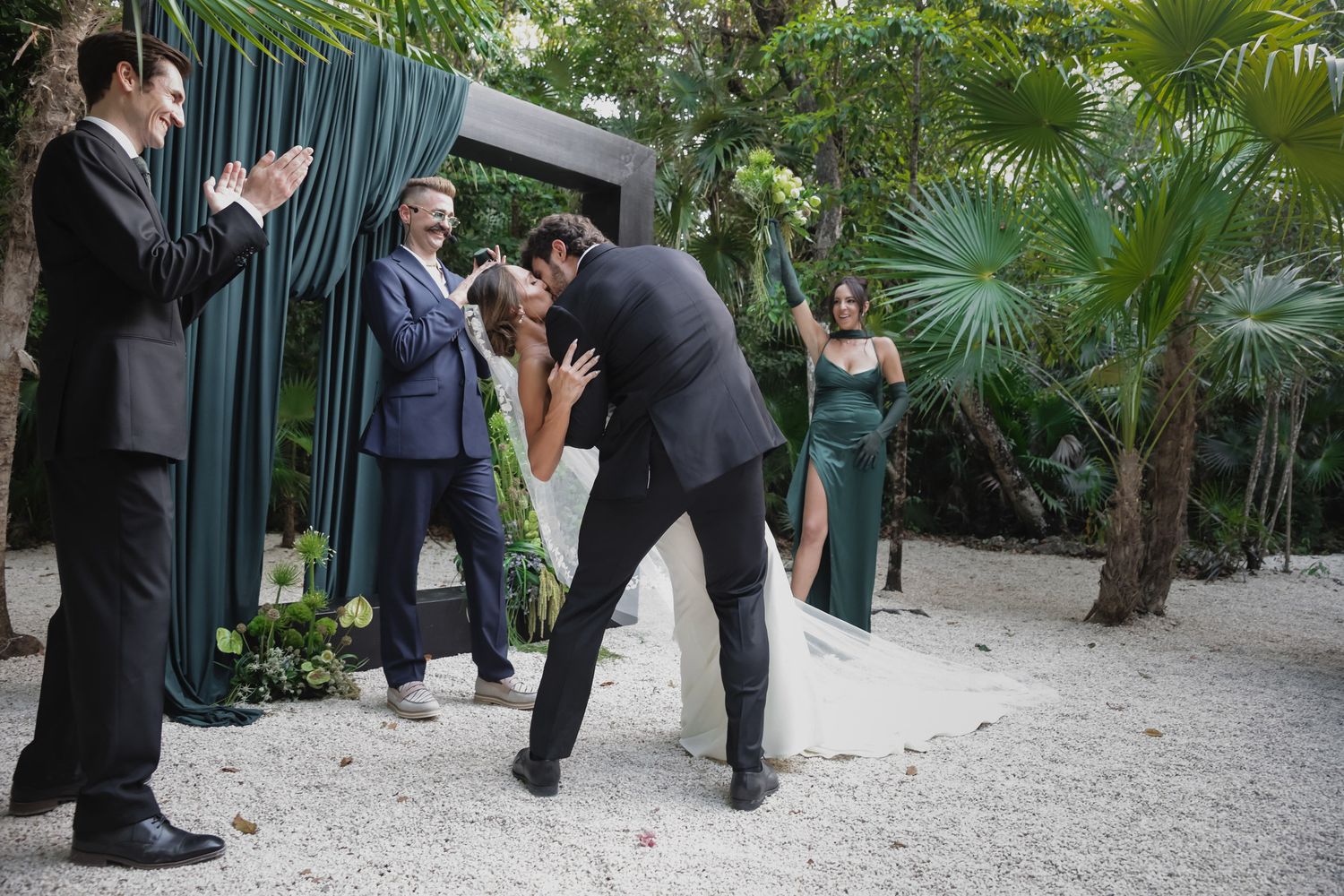 Groom dips bride for a kiss at outdoor tropical wedding ceremony while guests applaud near emerald draped arch.