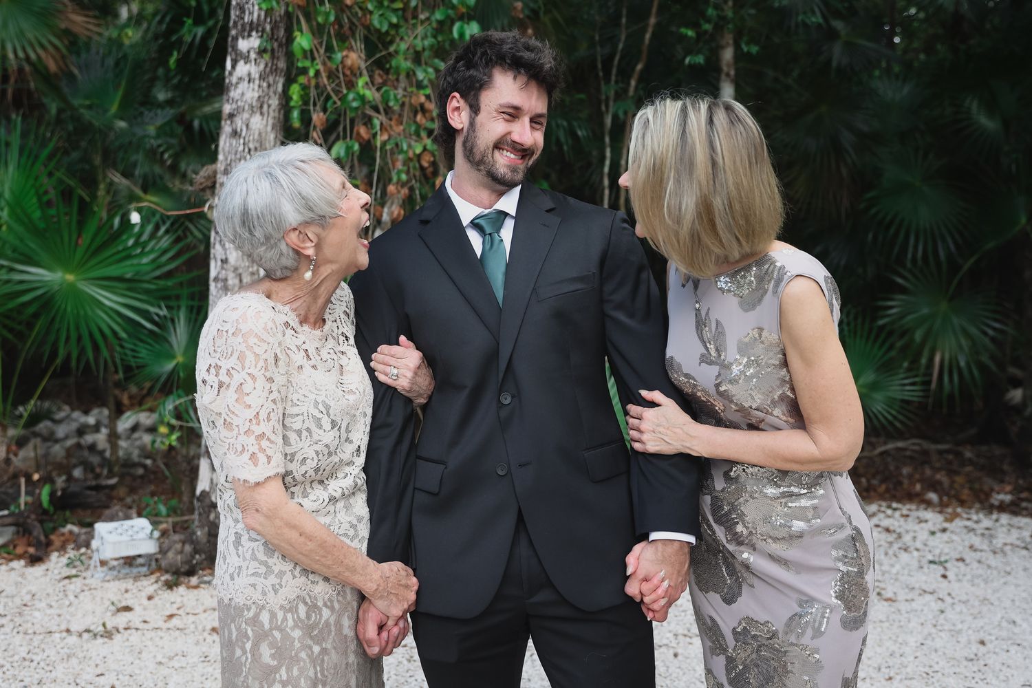 A groom in a black suit stands smiling between two elegant older women in formal dresses at an outdoor wedding.