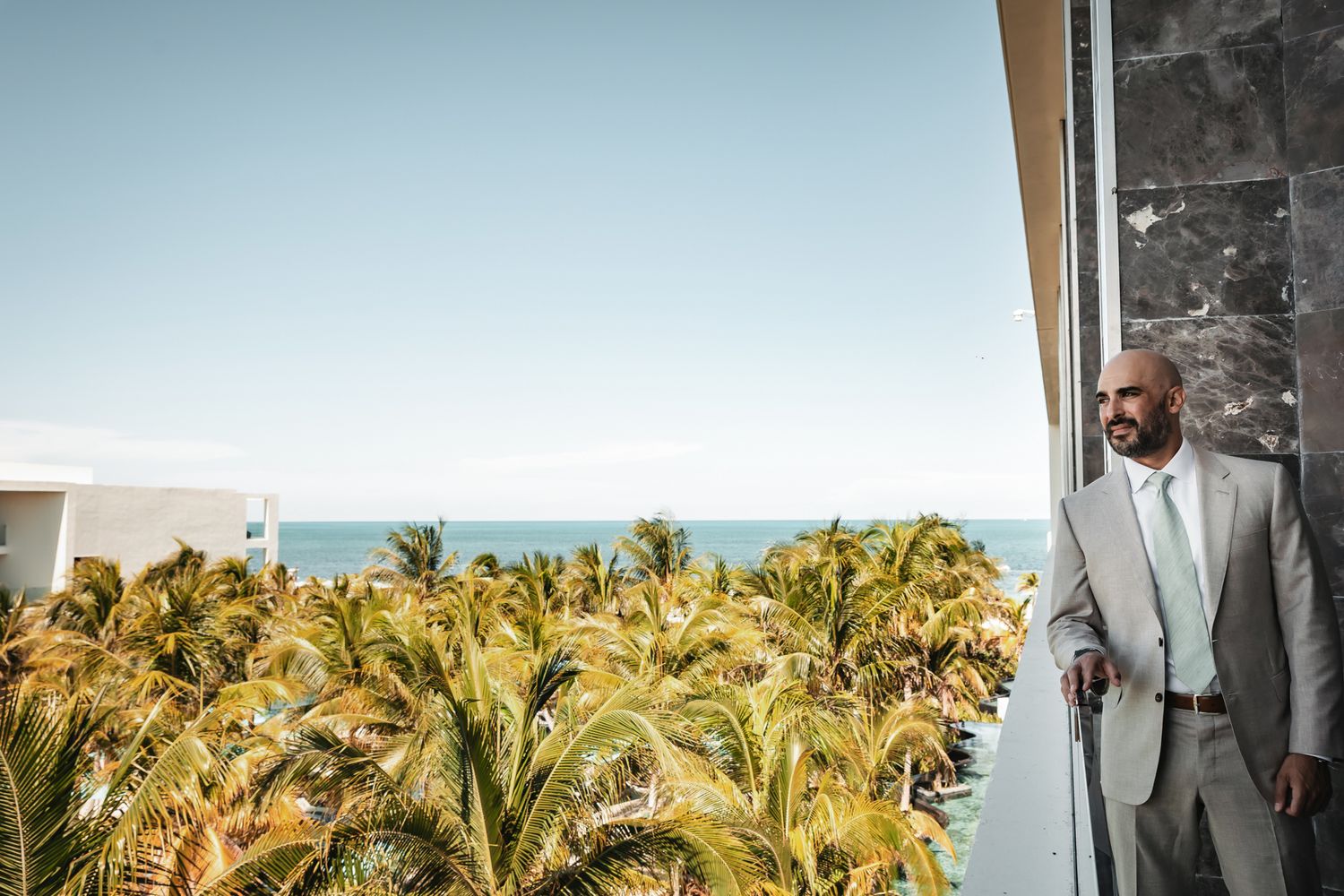 Elegant man in light gray suit stands on balcony overlooking tropical palm trees and ocean horizon.