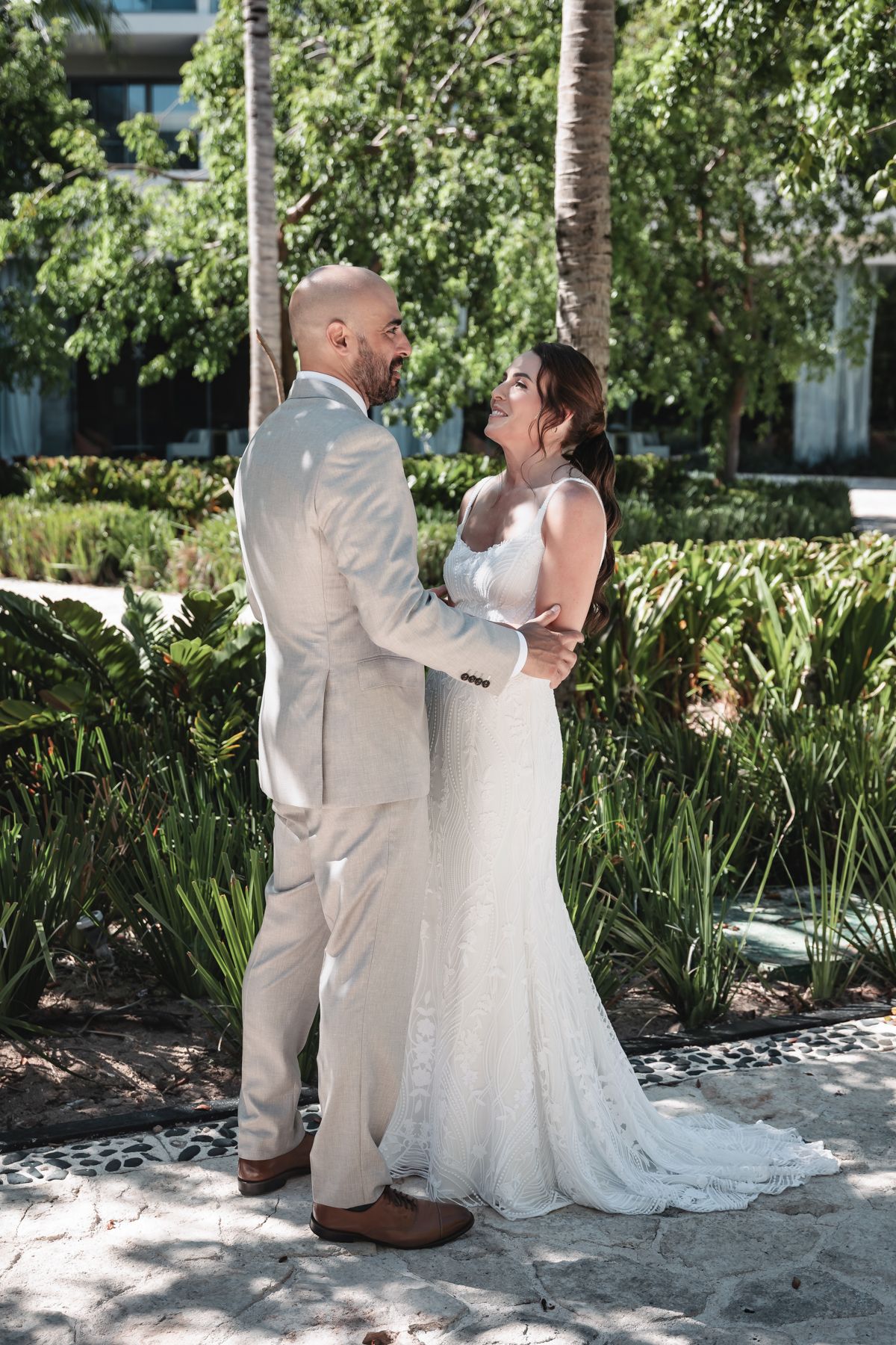 A bride and groom share a romantic embrace in a lush garden setting, with the groom in a beige suit and bride in a white lace gown.