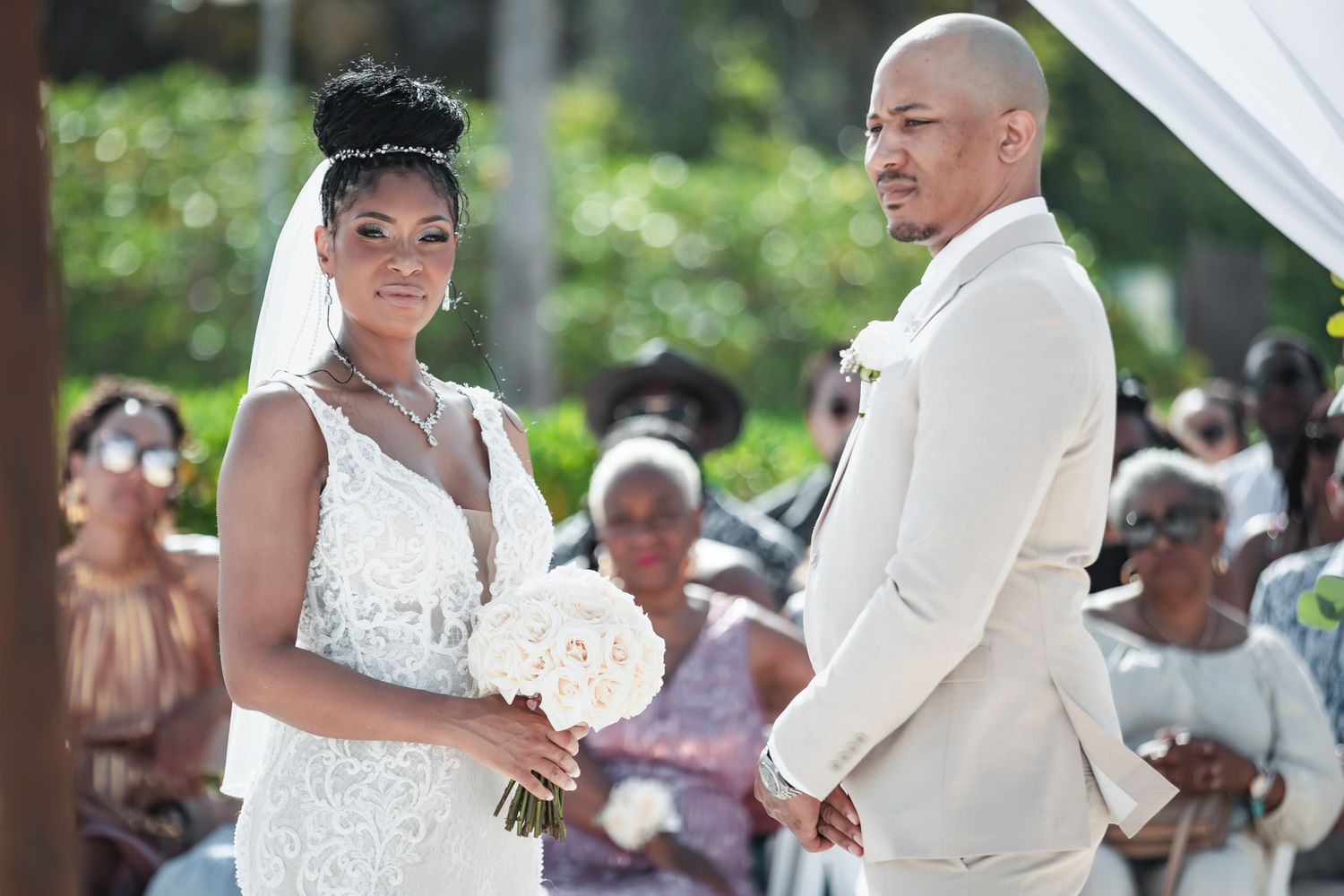 Bride and groom stand at outdoor wedding altar, bride in lace gown holding white bouquet, groom in cream suit.