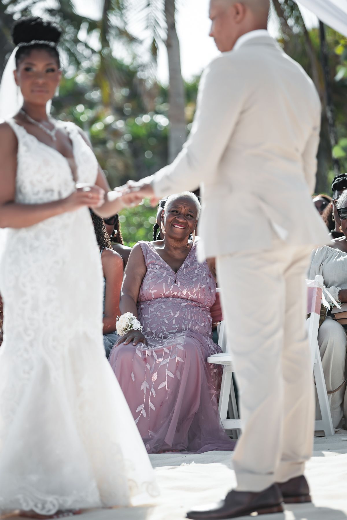 Bride and groom exchange rings at outdoor wedding ceremony while smiling guest in pink dress watches joyfully.