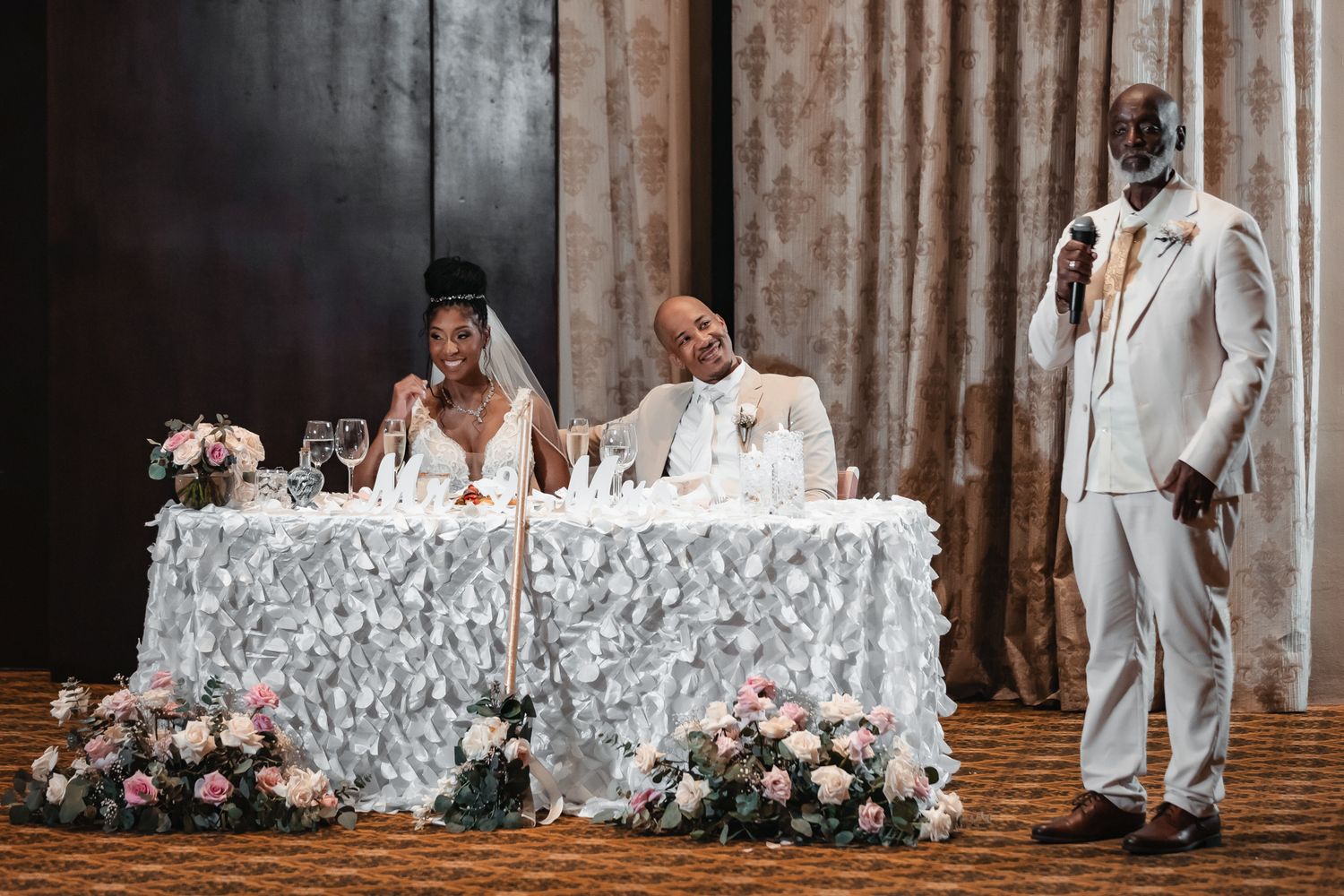 Bride and groom seated at sweetheart table during wedding reception while best man gives toast.