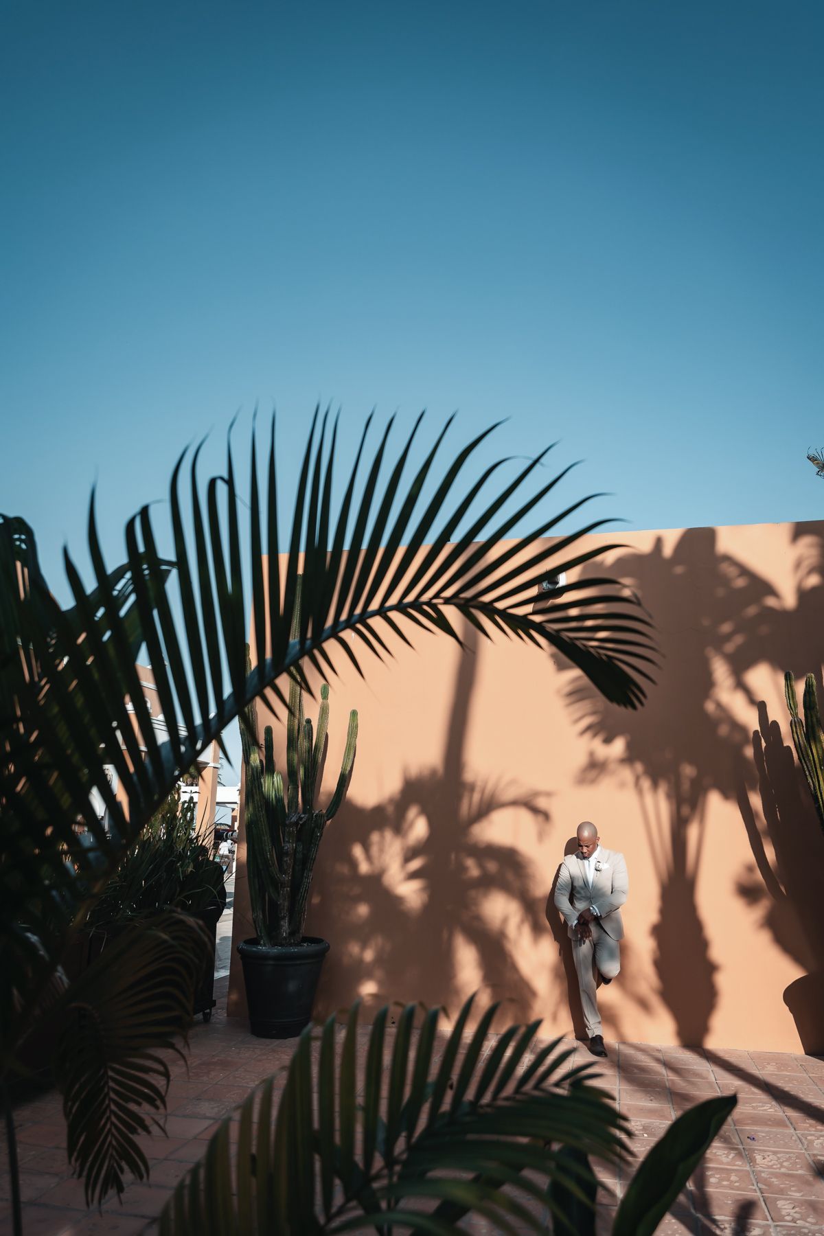 A man in a light suit walks beside a terracotta wall with palm tree shadows under a clear blue sky.