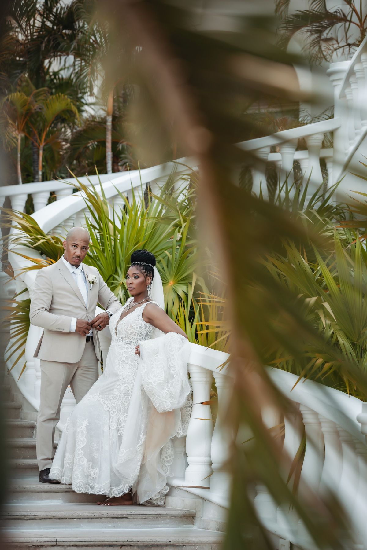 Bride in lace gown and groom in beige suit stand on white staircase framed by lush tropical foliage.