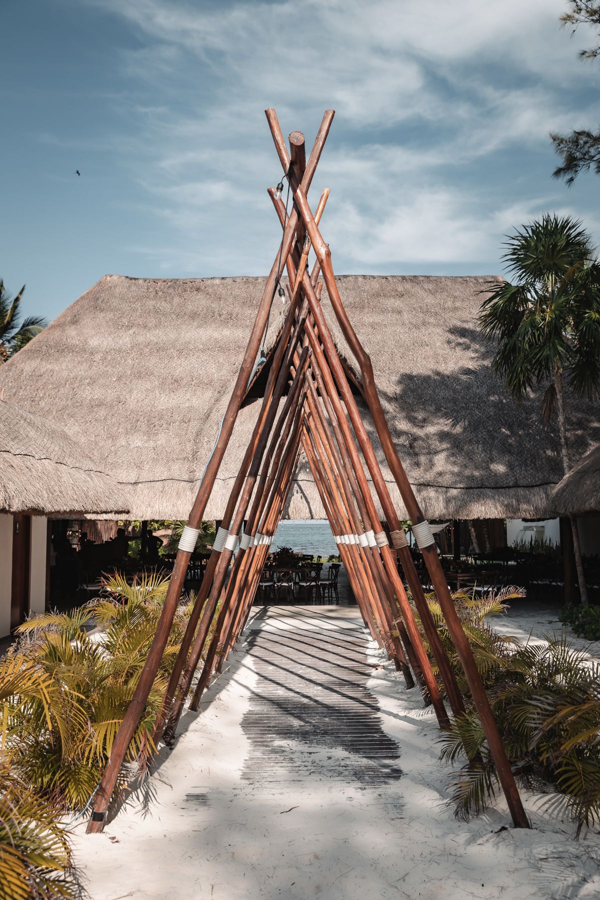 Rustic wooden teepee structure on white sand beach with thatched roof palapa and palm trees in tropical resort setting.