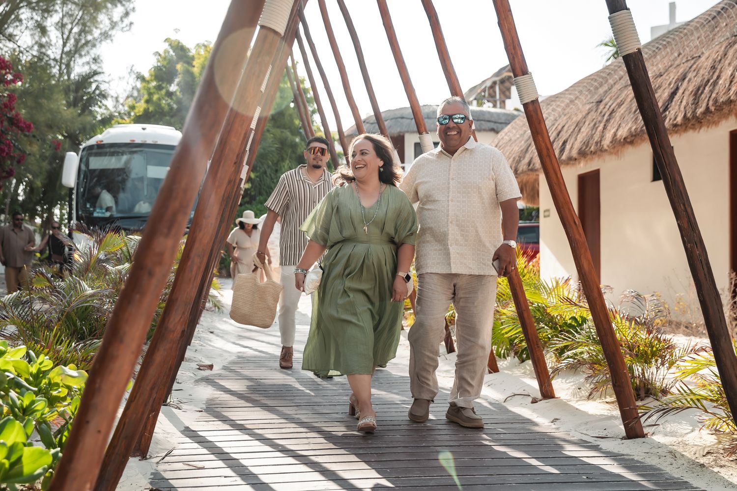Couple and friend walking through tropical resort pathway with thatched bungalows and lush greenery.