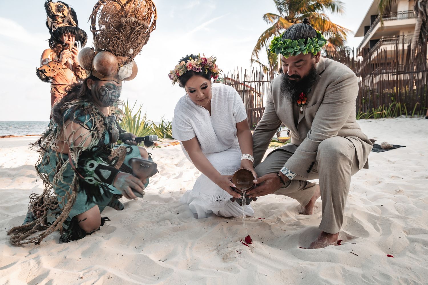 Couple exchanges vows on sandy beach with officiant in colorful costume during tropical destination wedding ceremony.