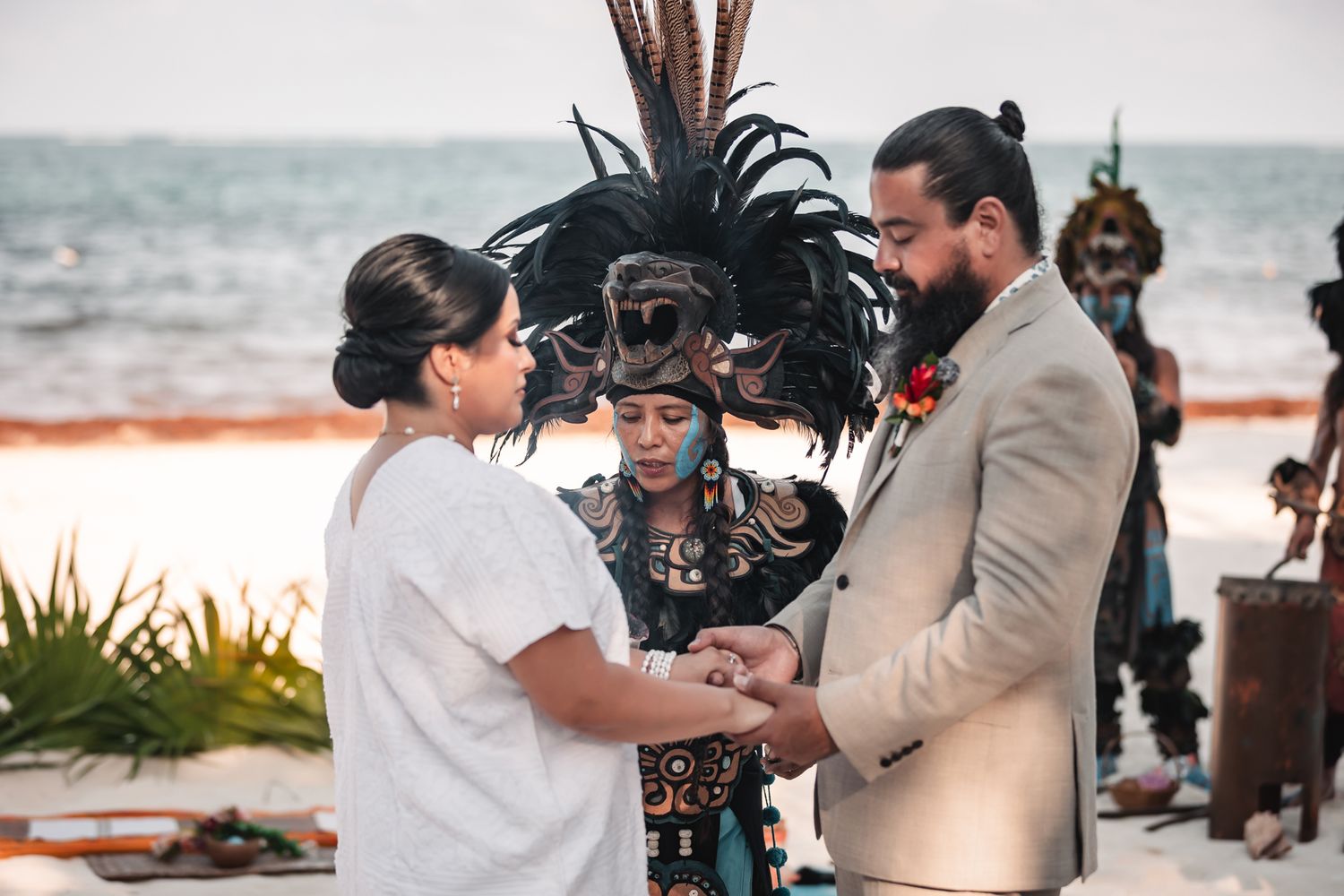 Couple exchanges vows at beach wedding with officiant wearing traditional Mayan headdress and attire by the ocean.