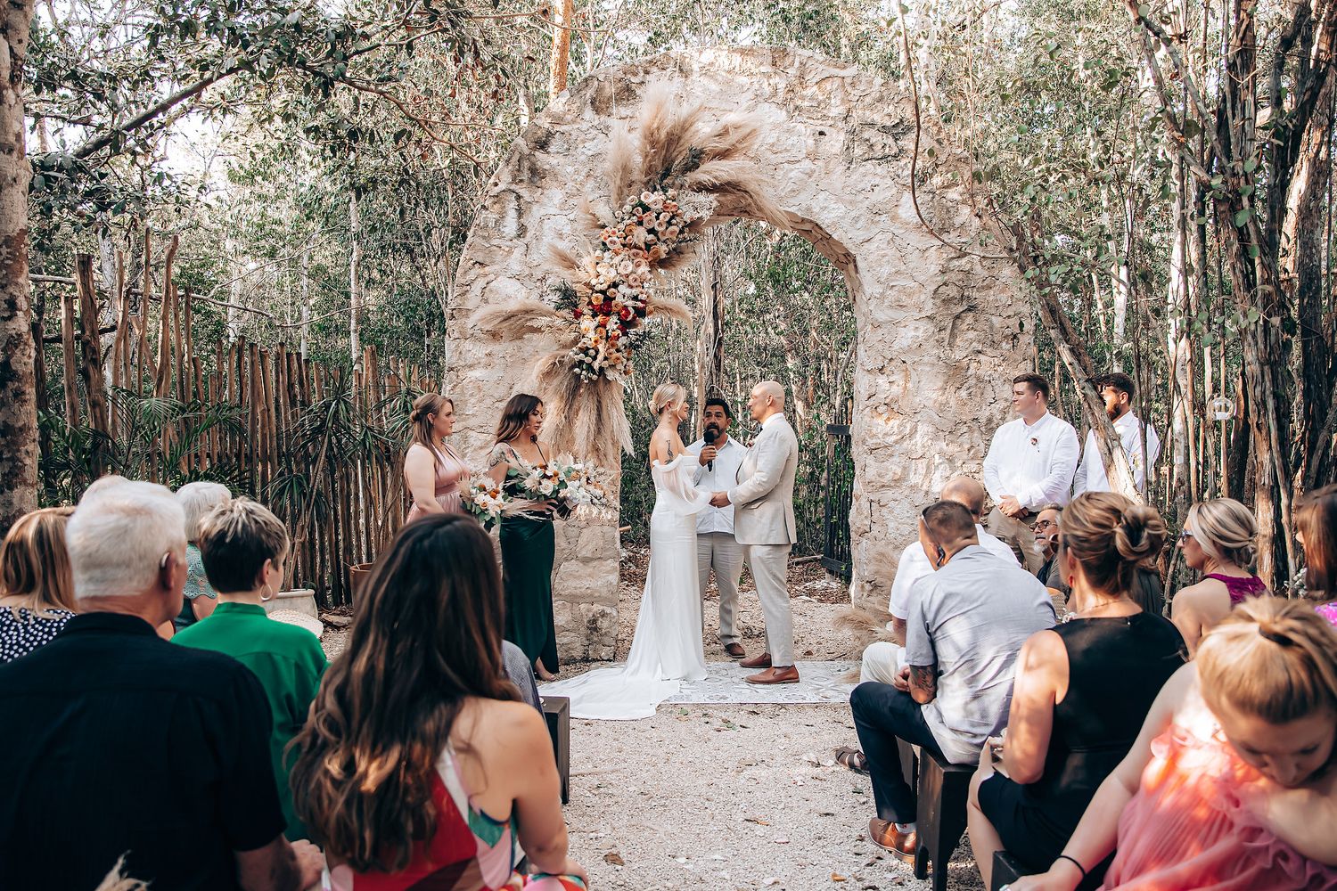 Outdoor wedding ceremony in a natural wooded setting with guests seated on wooden chairs.