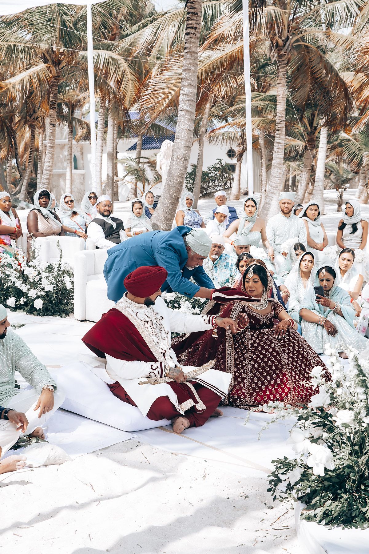 Wedding guests seated on white cushions under palm trees at a beachside ceremony.