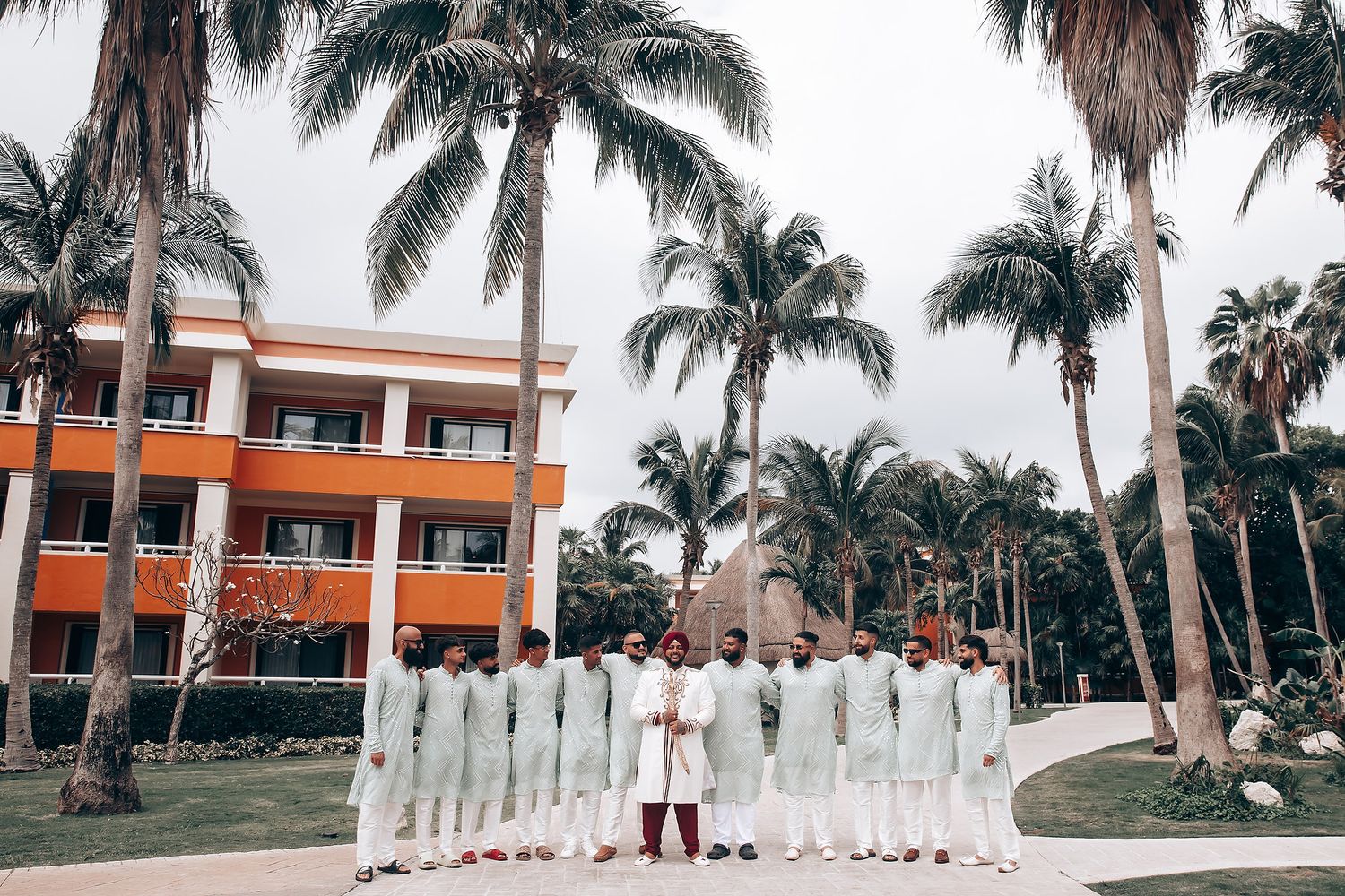 Wedding party in white attire standing with palm trees and orange building in background.