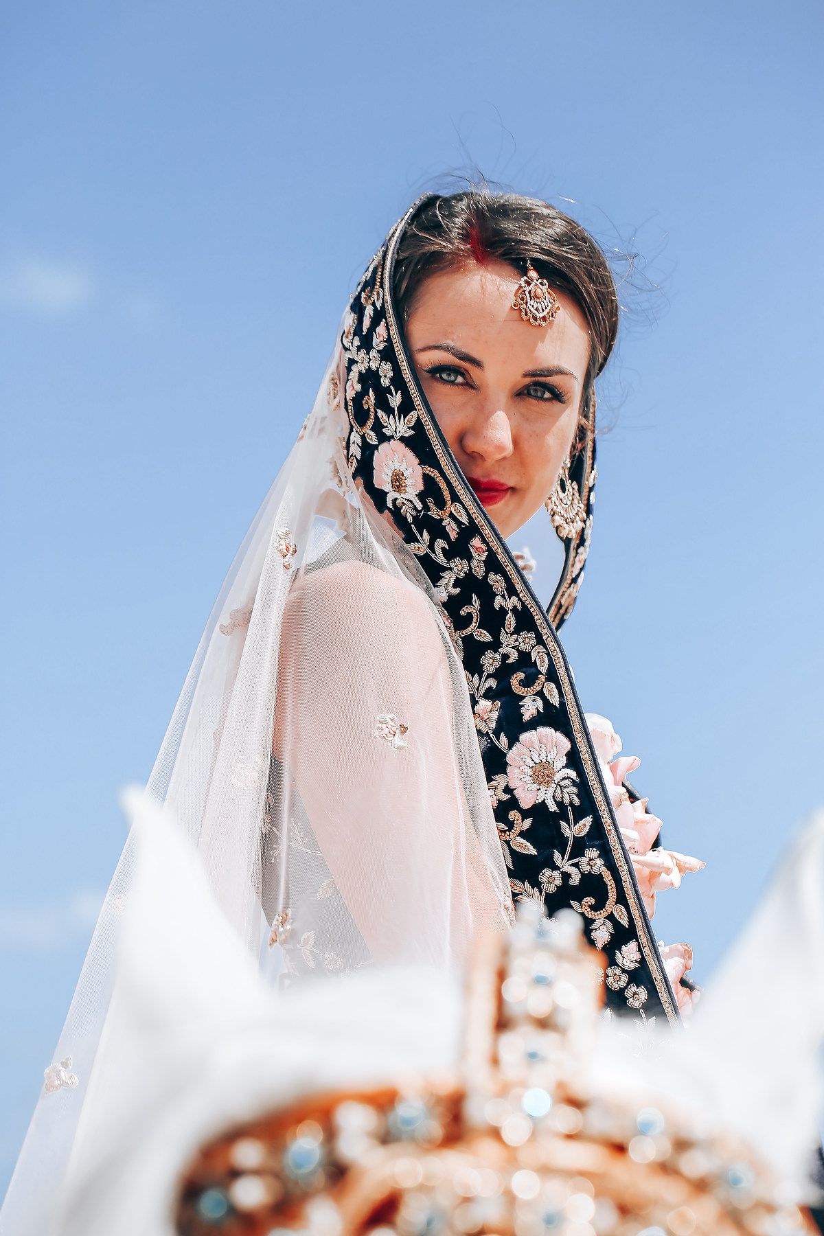 Person wearing black and white floral headscarf poses against bright blue sky.